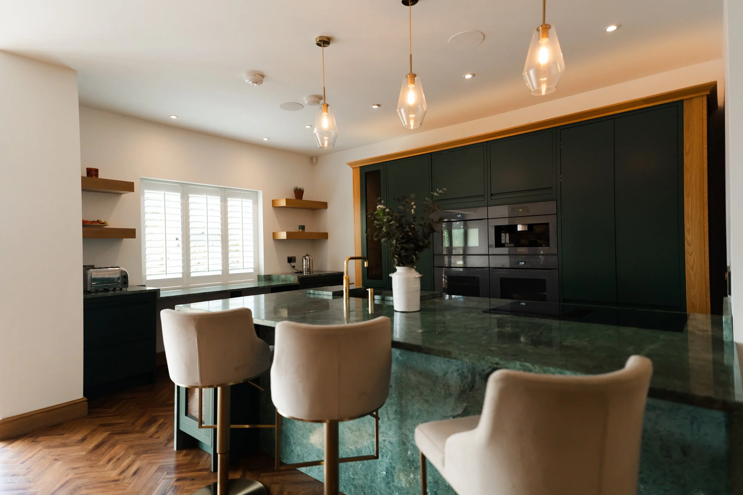 A modern kitchen with green cabinets, a marble island with a vase of flowers, pendant lights above, and a window with white shutters.