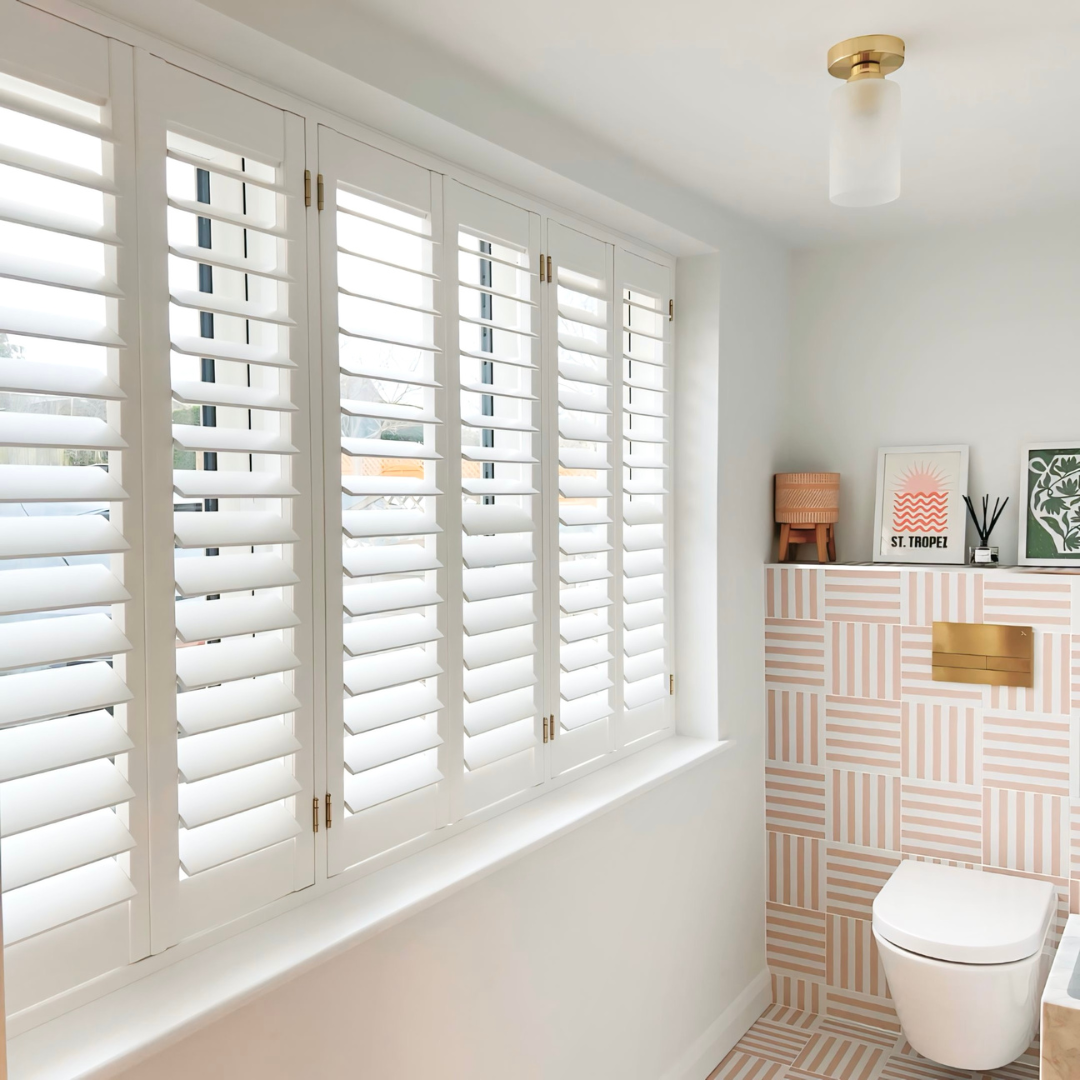 Bright bathroom with white plantation shutters on the window, pink and beige geometric patterned tiles on the wall and floor, a white toilet, and decorative items on a shelf including framed artwork, a wooden lamp, and diffuser sticks.