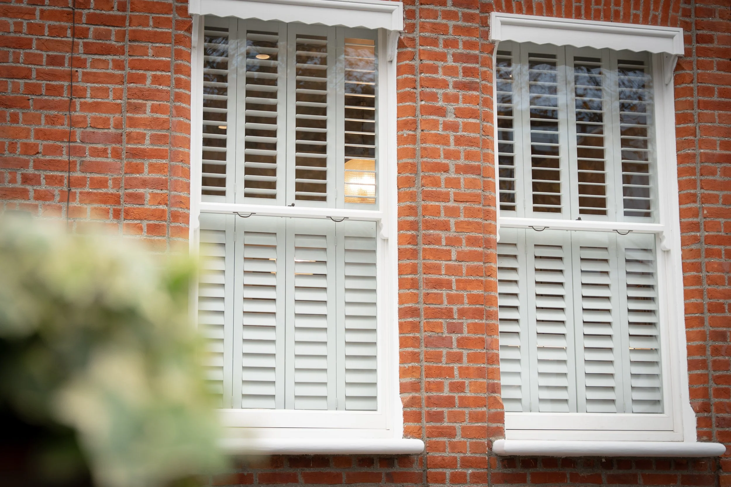 Close-up of a red brick house exterior with two white-framed windows with plantation shutters and a small overhang on top of each window.
