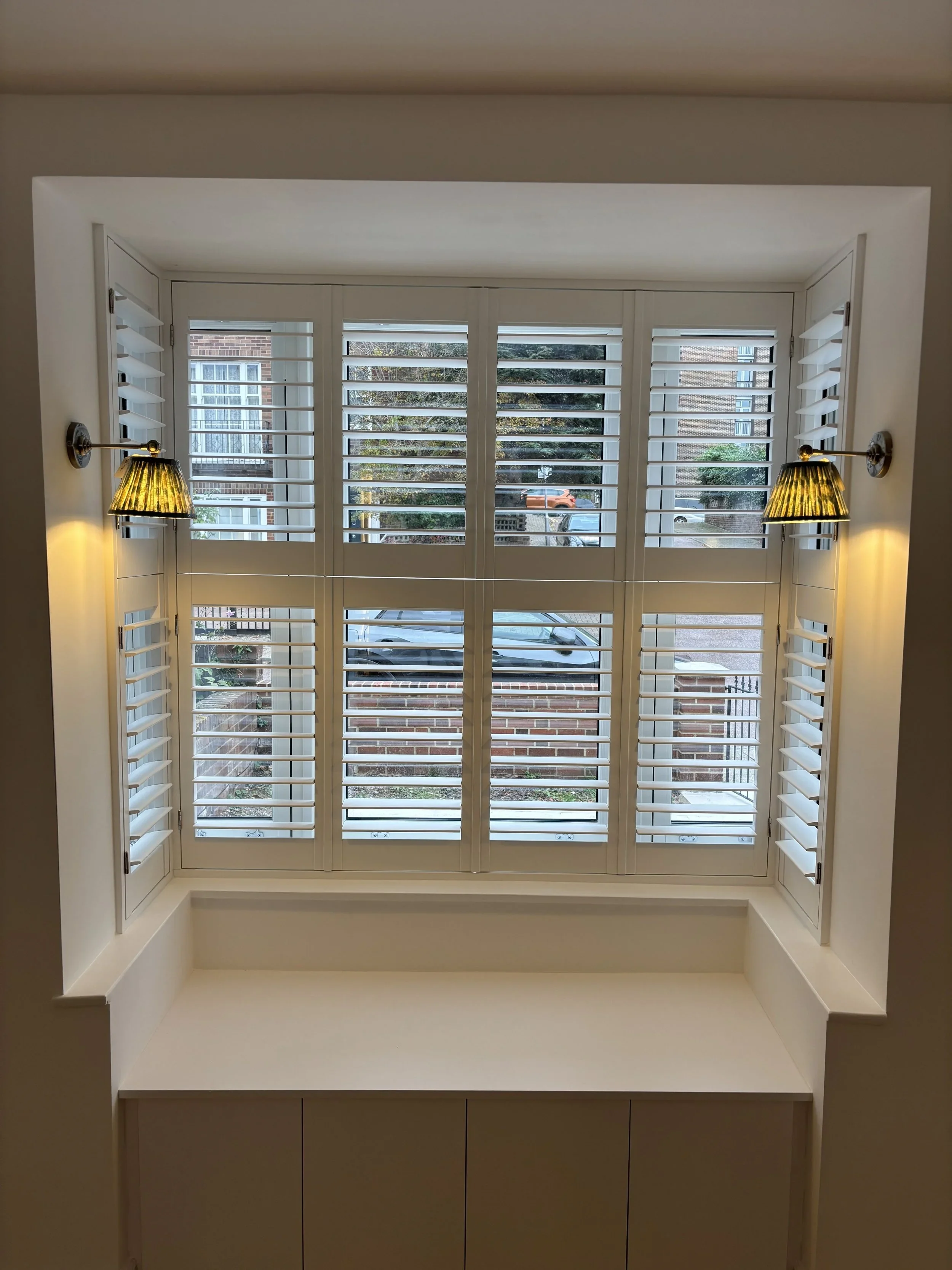 Interior view of a window with white plantation shutters flanked by wall-mounted lamps with yellowish shades.