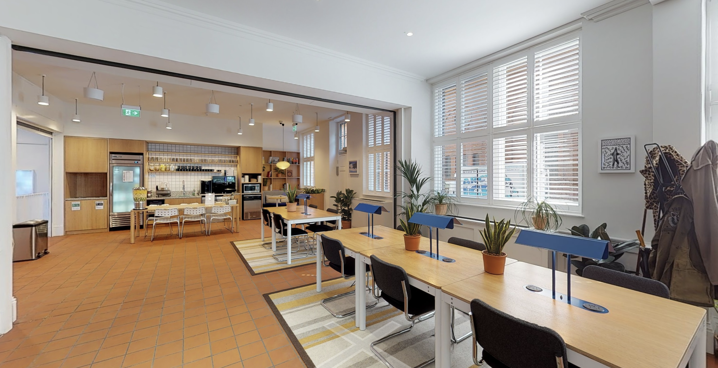 Interior of a bright, modern communal room with large windows, several wooden tables with black chairs, indoor plants, and a kitchenette area with wooden cabinets, a microwave, and a few chairs. Cluttered clothing and coat hooks are visible on the ri