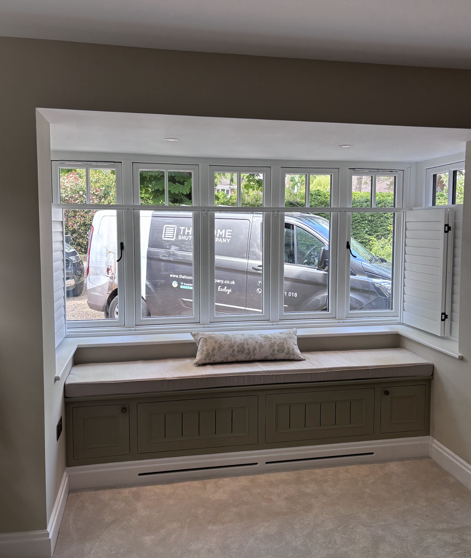 A large window seat with a beige cushion and a decorative pillow, situated under a wide bay window with white frames and open shutters, overlooking a driveway with parked vehicles and greenery outside.