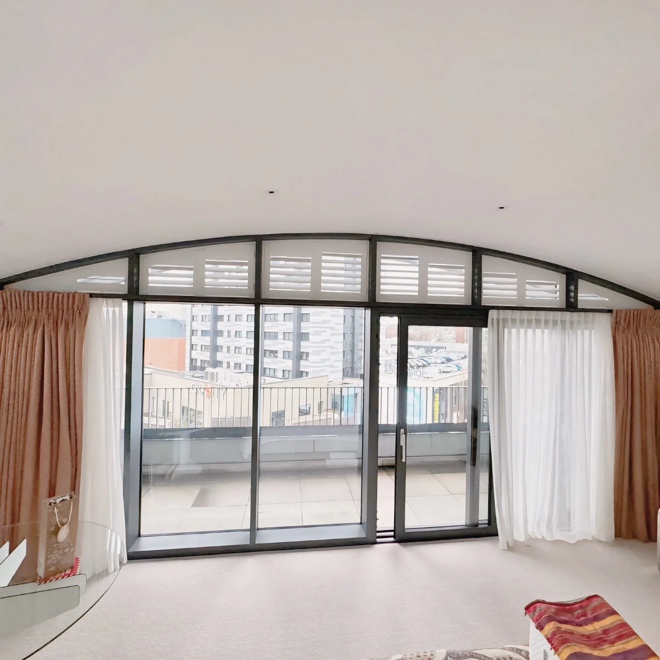 Living room with large window, curtains, and sliding glass door leading outside, with buildings visible in the background.