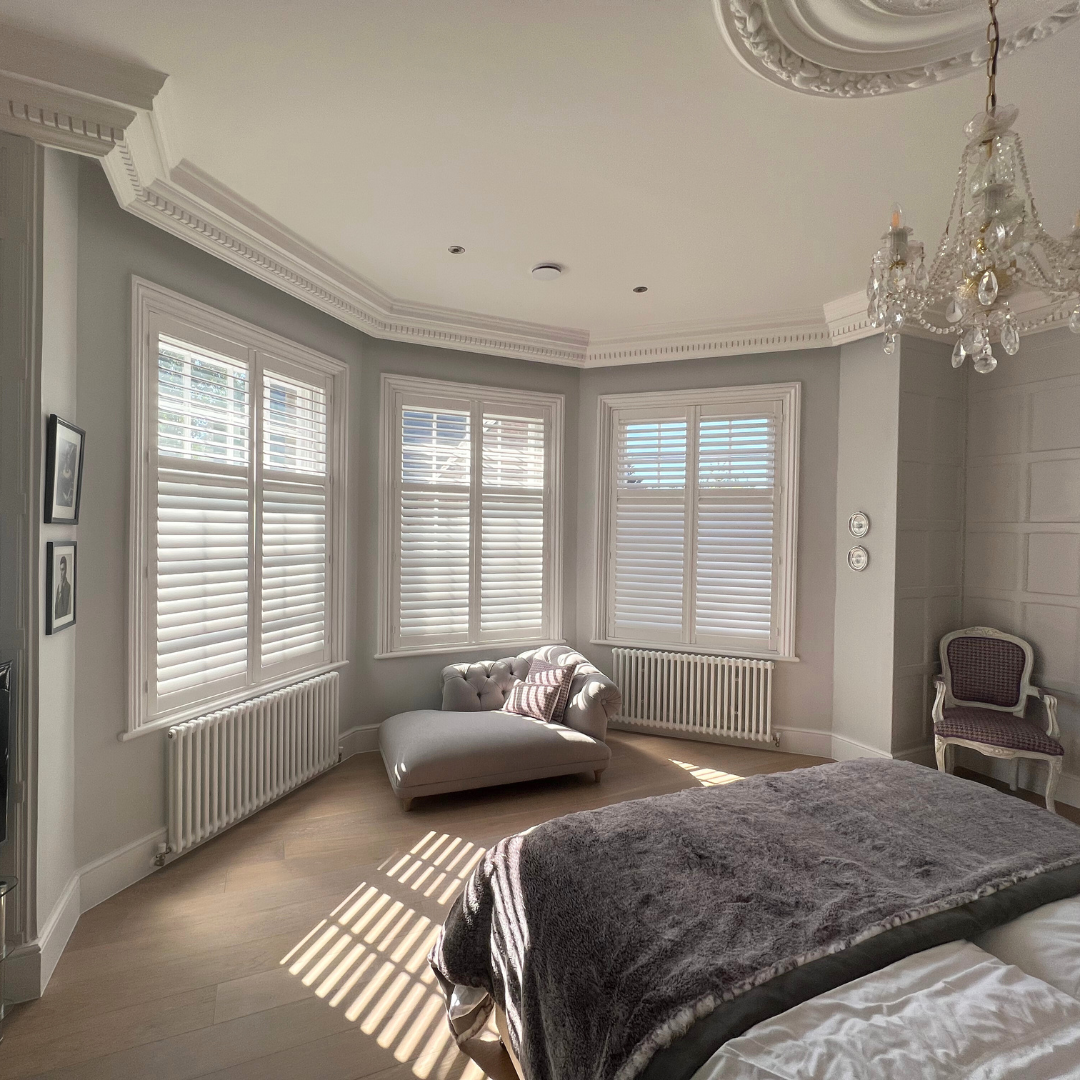 Bright bedroom with large windows, white shutters, a tufted lounge chair, a bed with a plush blanket, and elegant decor including a chandelier.