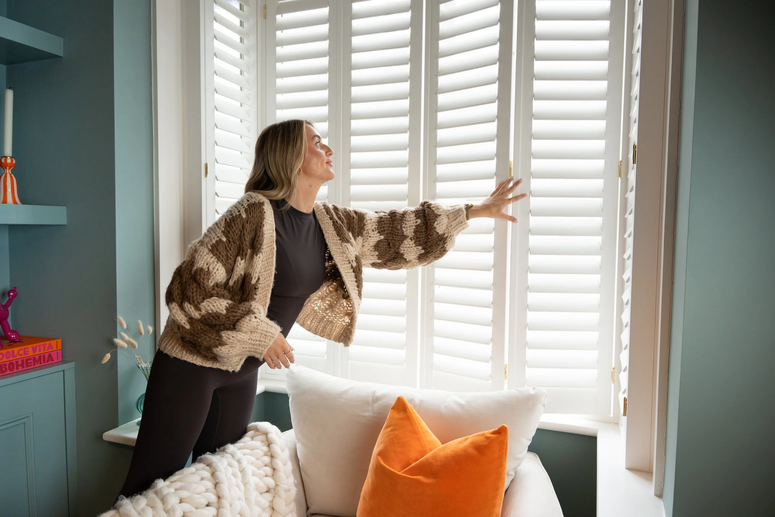 A woman looking out the window with white shutters, wearing a brown and beige cardigan and black pants, in a room with teal walls and decorative items.