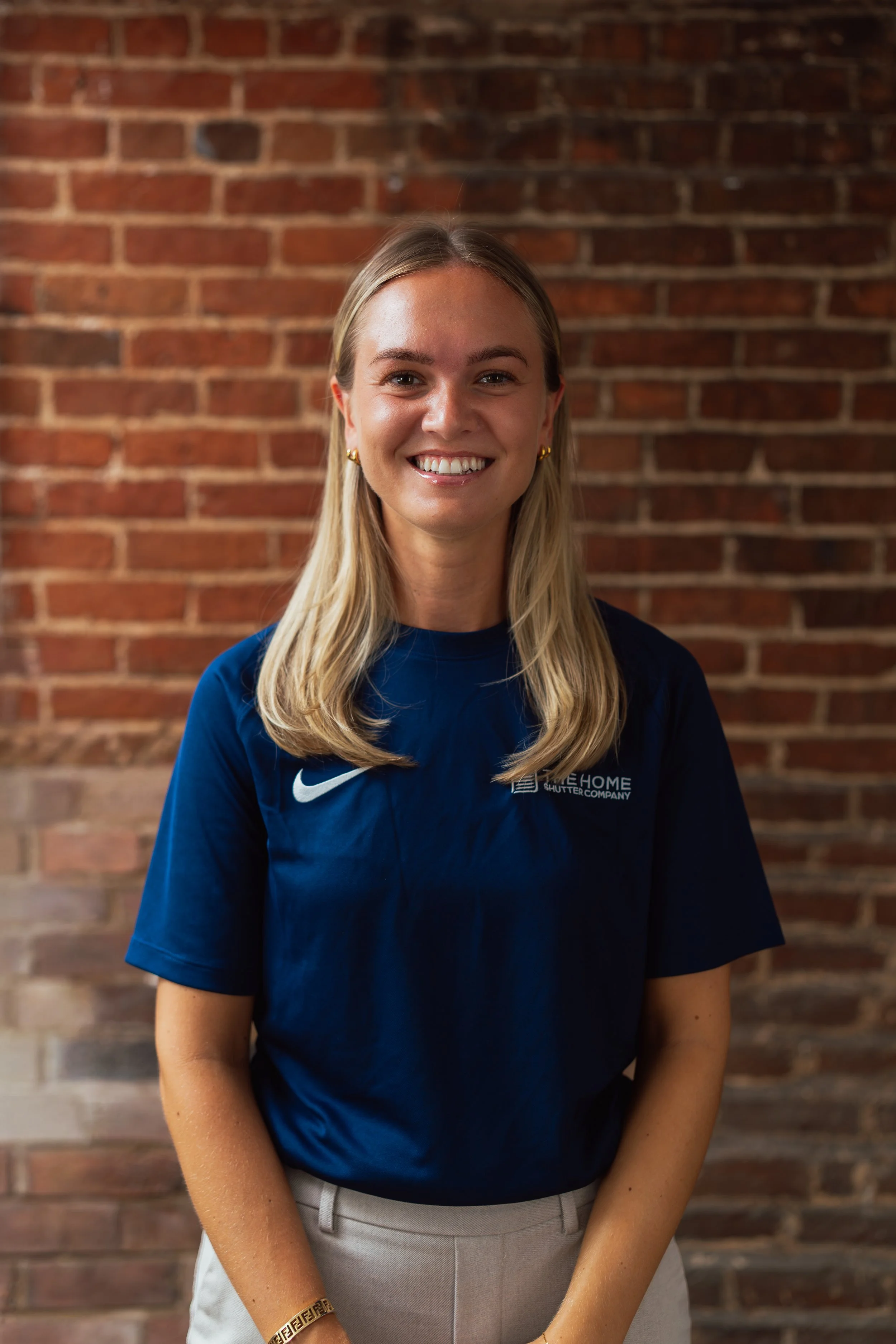 A young woman with blonde hair smiling, wearing a navy blue Nike shirt with a logo and text, standing in front of a brick wall.