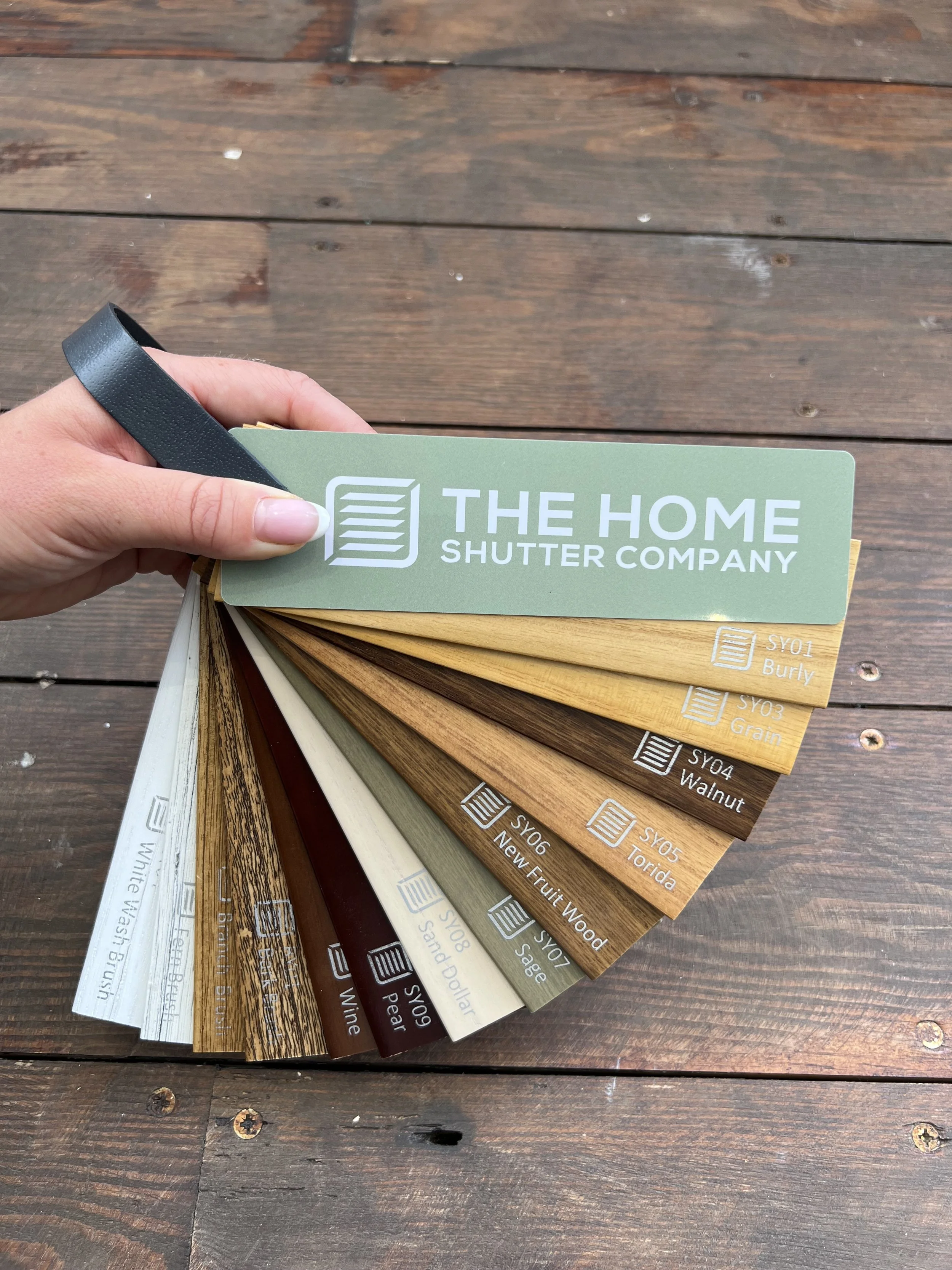 A hand holding a fan deck of wood stain color samples from The Home Shutter Company, fanned out to show various wood finish options on a wooden surface.
