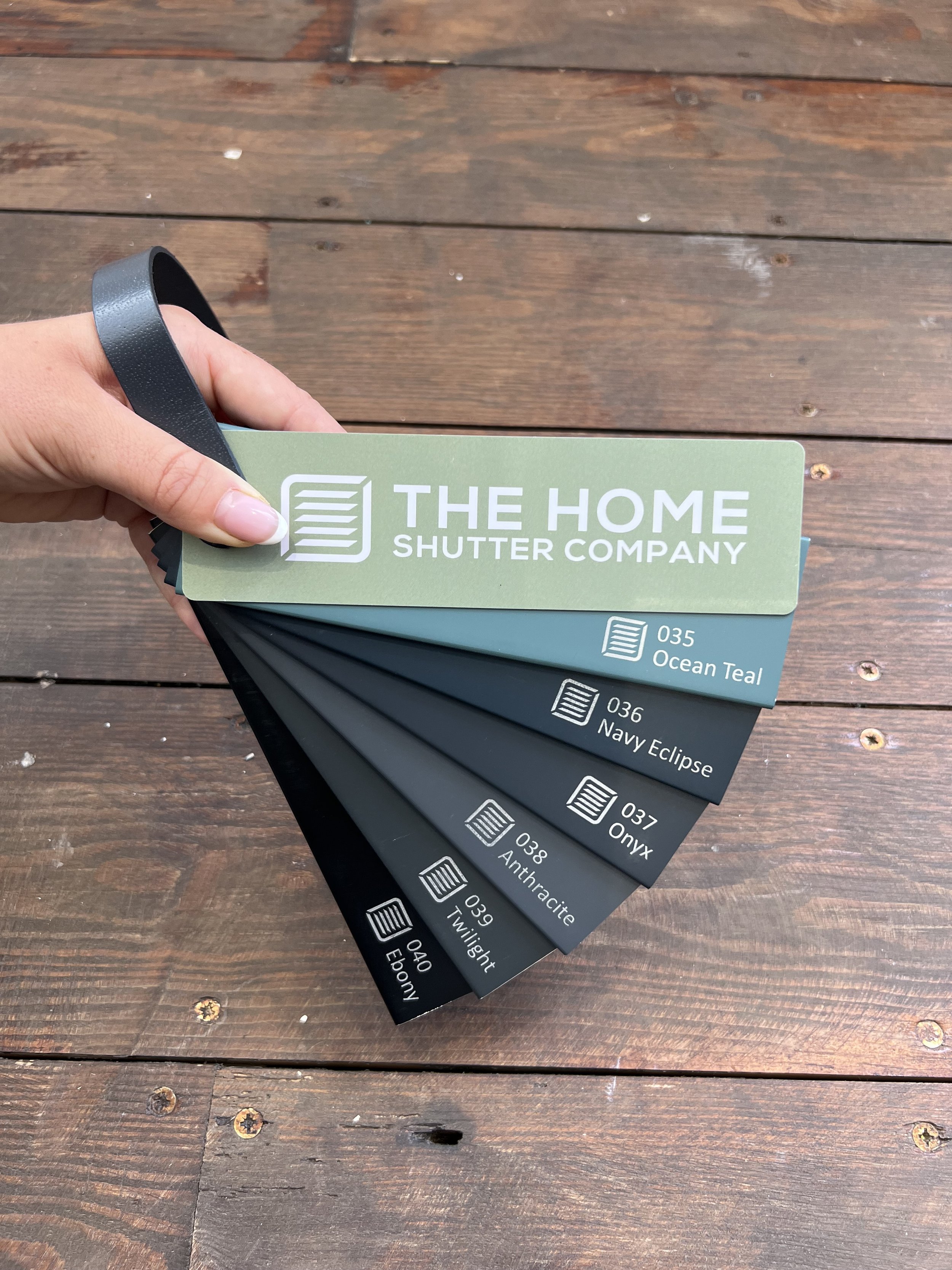 A person's hand holding a color swatch sample fan deck from The Home Shutter Company, with multiple color options visible, including Ocean Teal, Navy Eclipse, and Onyx, against a wooden surface.
