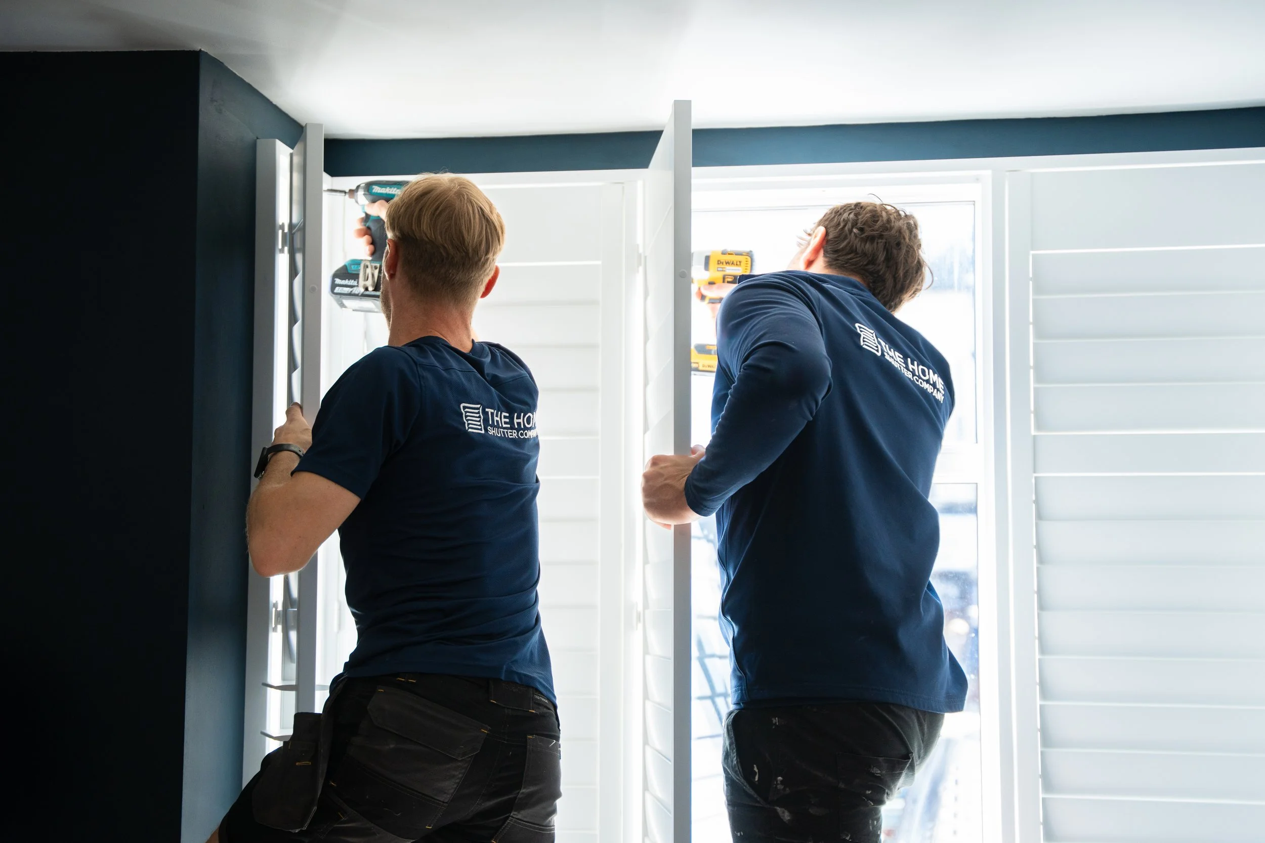 Two men installing window shutters, one using a power drill, both wearing navy shirts that read 'The Home Shutter Company', inside a bright room with white shutters and sunlight.
