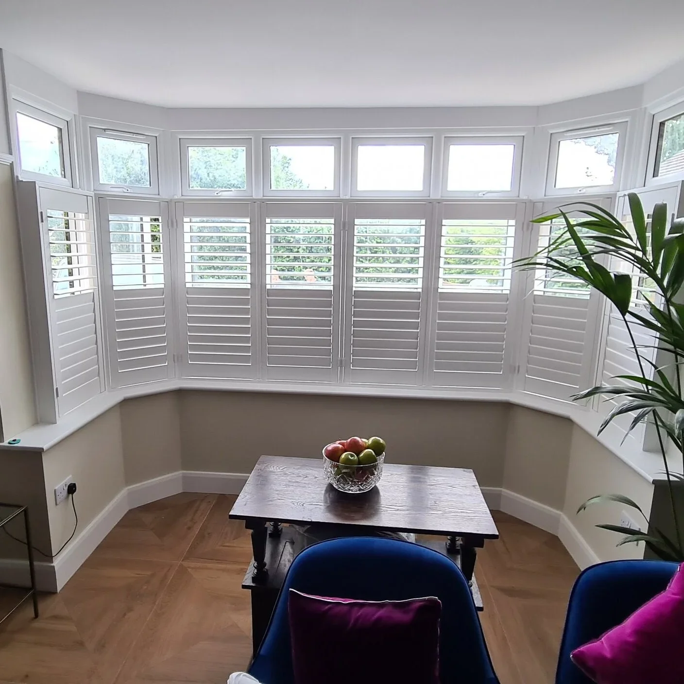 Bright dining area with a large bay window featuring white shutters, a wooden table with a bowl of fruit, and blue chairs with purple cushions.
