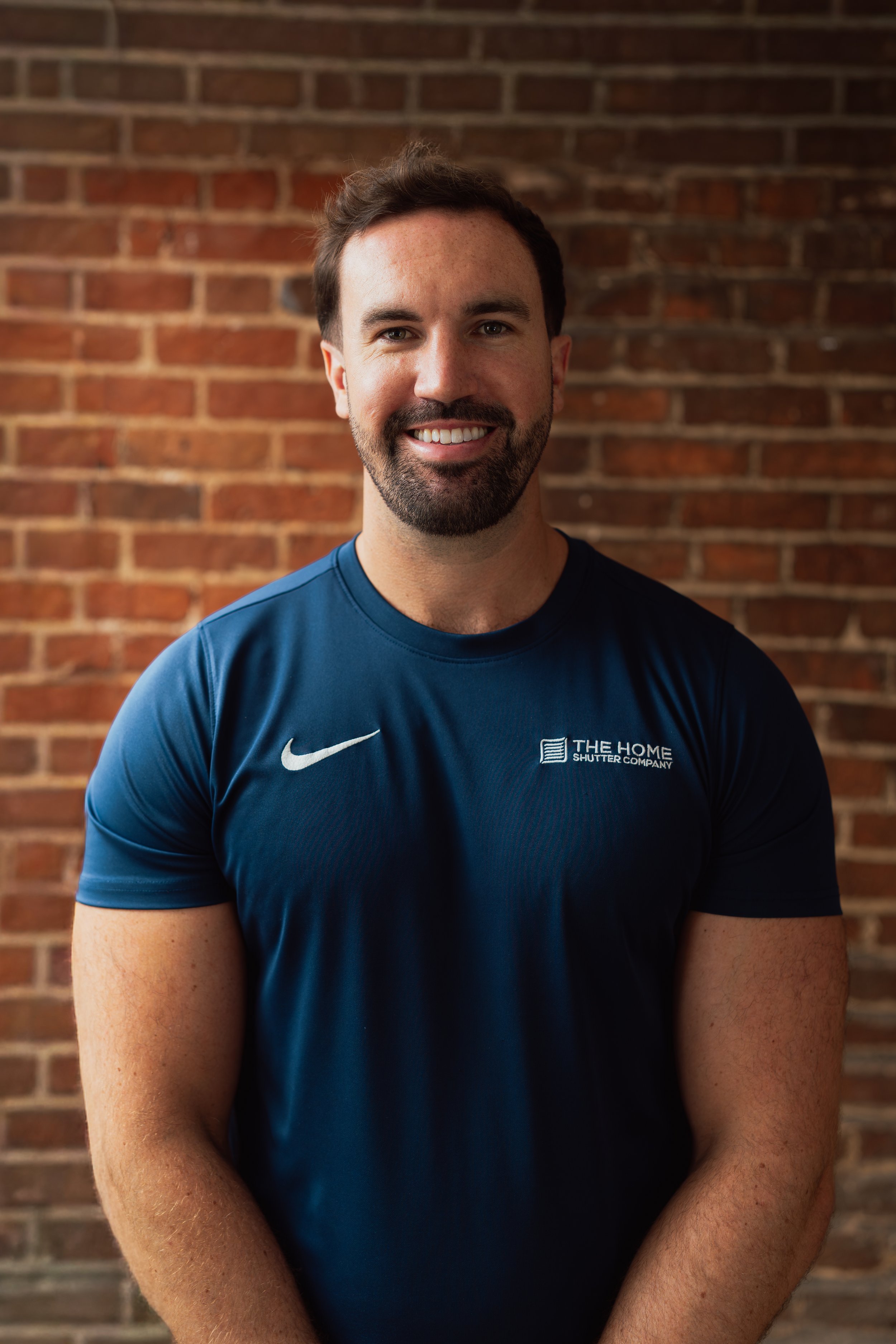 A smiling man with short brown hair, beard, and blue eyes, wearing a navy blue Nike t-shirt with a logo and text printed on it, standing in front of a red brick wall.