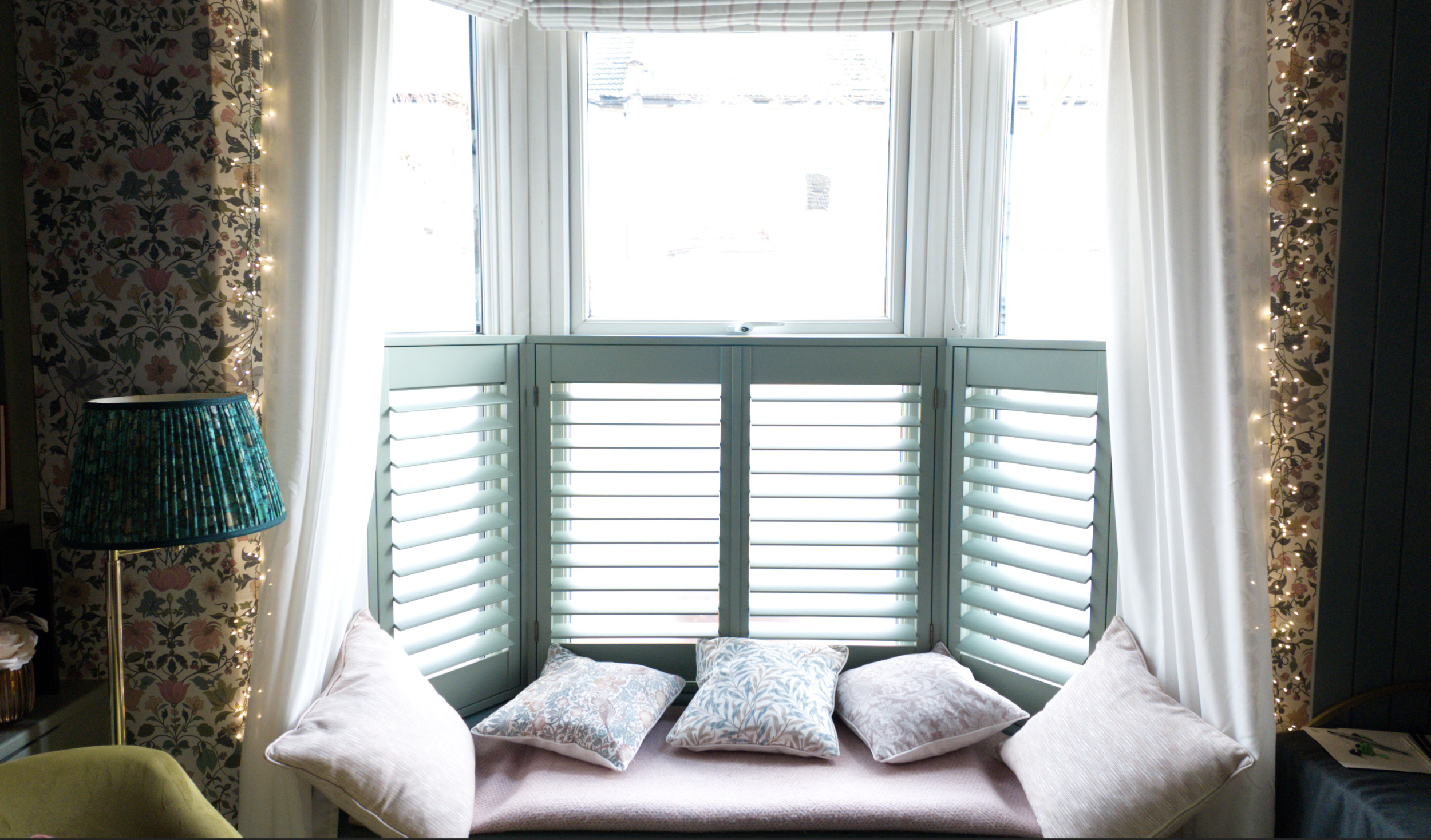 A cozy sitting nook with pastel cushions and a window with white shutters, framed by floral and plaid curtains, decorated with string lights and a turquoise lampshade.