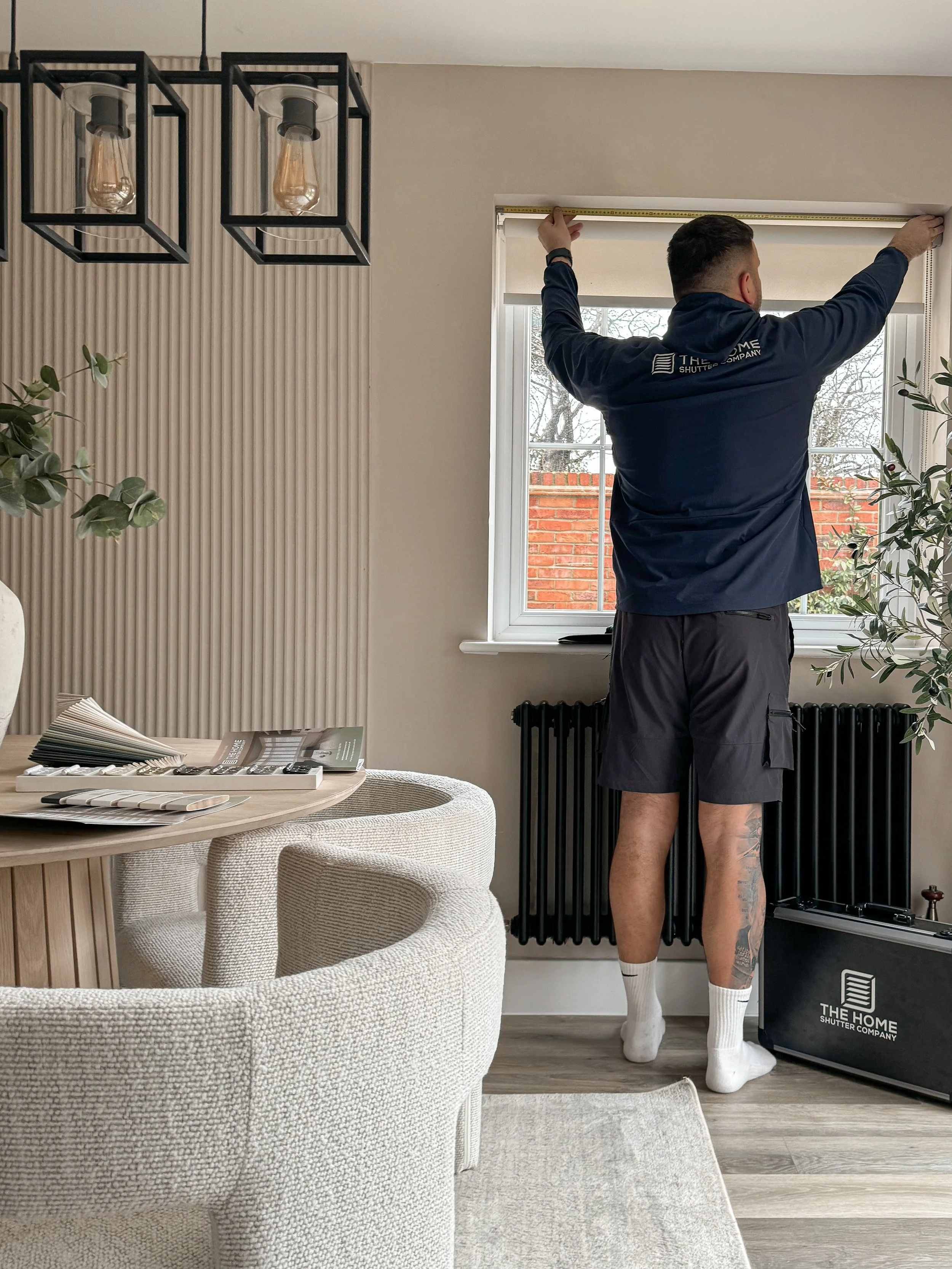 Man standing on a mat, measuring the window width with a tape measure in a modern living room, with a beige textured couch and botanical plants, and a ceiling light fixture.