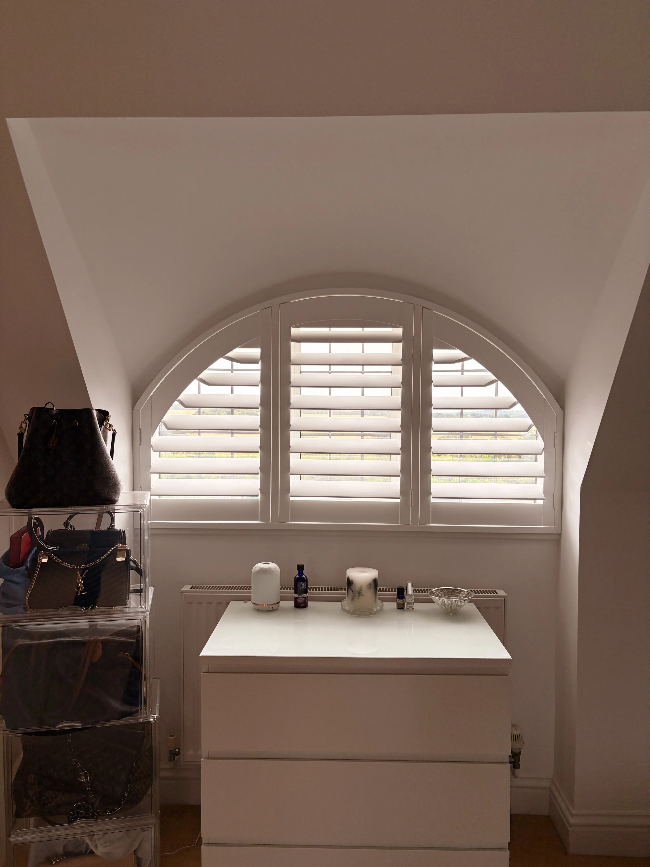 A room with a large arched window with white shutters. There is a white table with various items including candles and bottles. To the left, a black purse sits on a transparent storage unit.