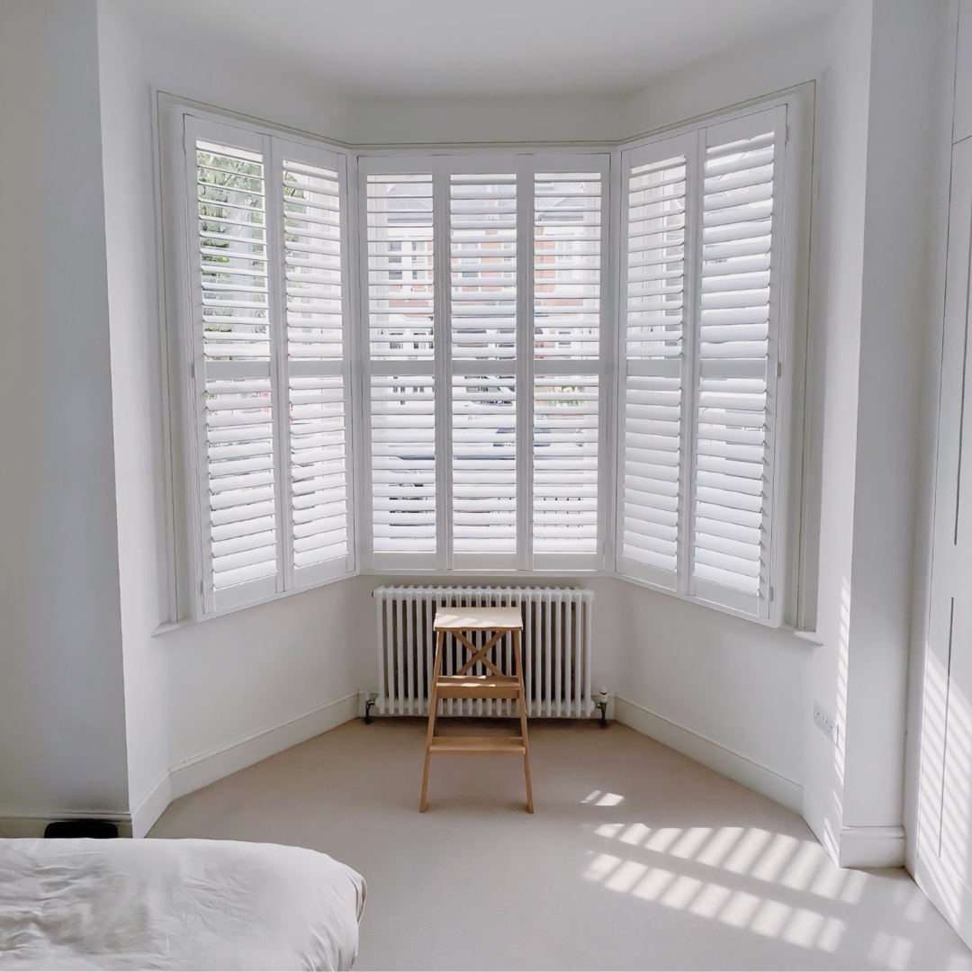Bright room with white walls and a large bay window with white plantation shutters. There is a small wooden chair in front of a white radiator underneath the window, and the corner of a bed with white bedding is visible in the lower left corner.