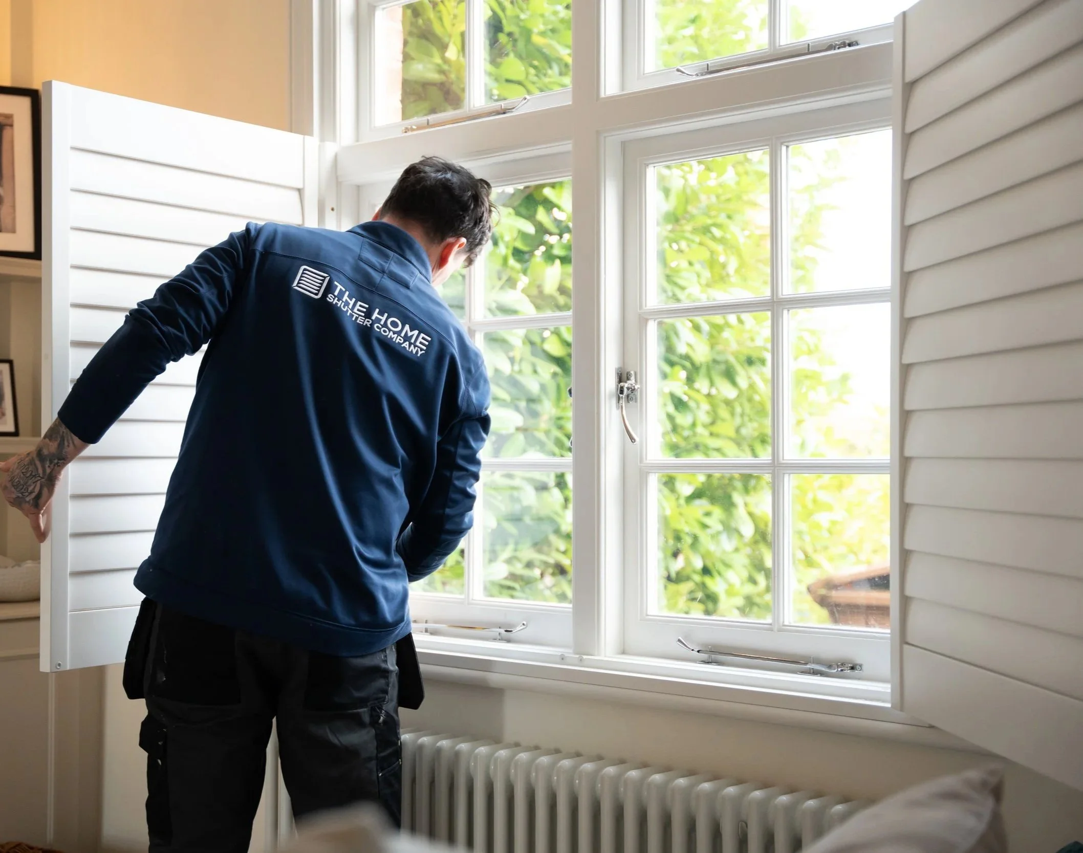 A worker in a blue uniform labeled 'The Home Shutter Company' installing or inspecting a white window shutter inside a house, with a view of green foliage outside the window.