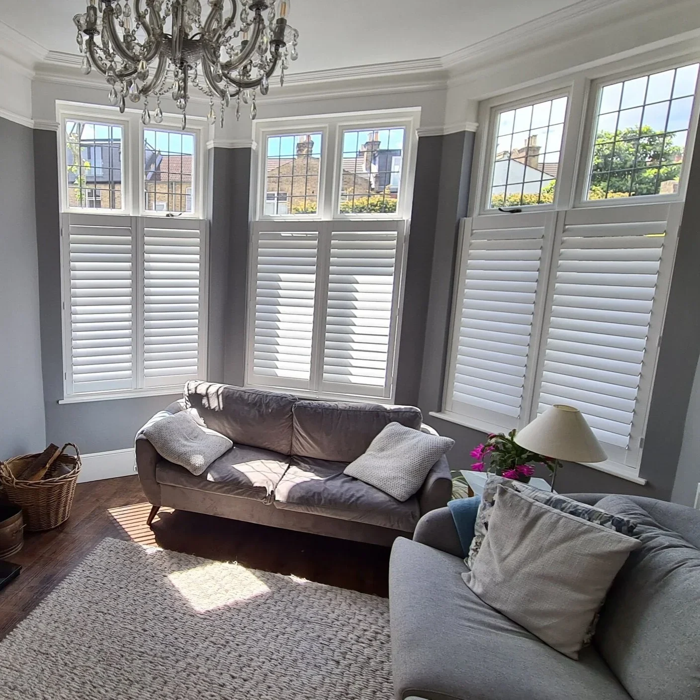 Living room with large bay window with white shutters, light grey walls, grey sofas, and a chandelier.