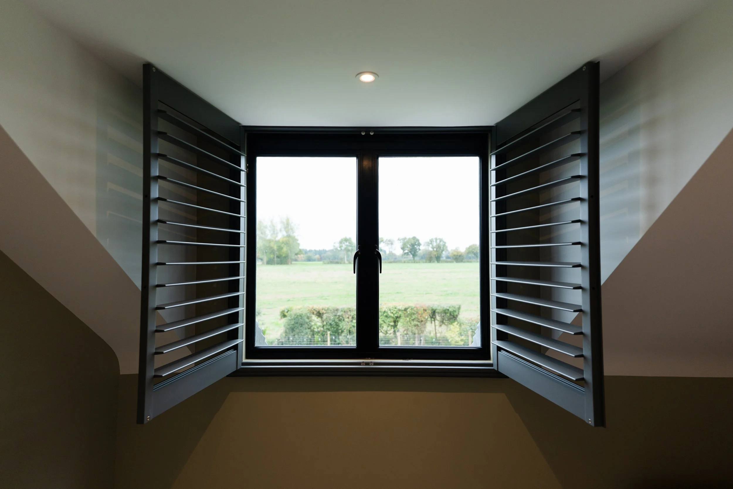 View through a large window with black frames, showing a green landscape with trees and grass, partially obscured by window shutters on both sides.