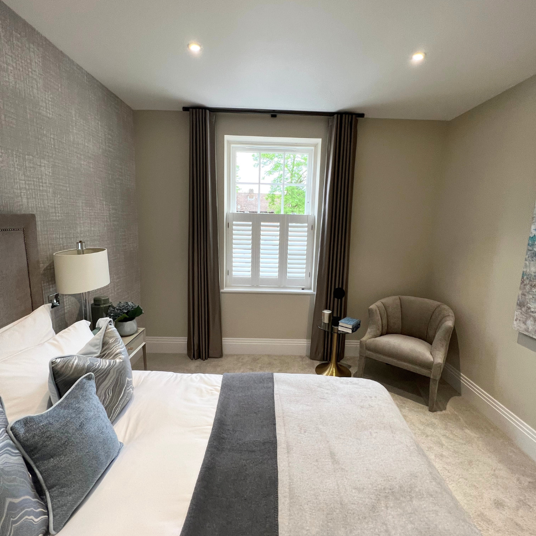 A bedroom with a bed, pillows, a nightstand with a lamp, a window with shutters and curtains, a small round side table with books, a cushioned armchair, and artwork on the wall.