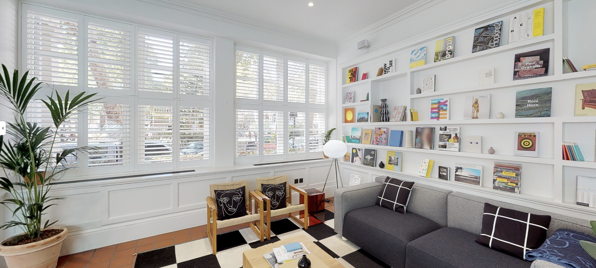 Living room with large white-walled windows with blinds, white bookshelves filled with books and art, a gray sofa with cushions, two wooden chairs with black face art cushions, a white floor lamp, potted green plant, and a black and white patterned r
