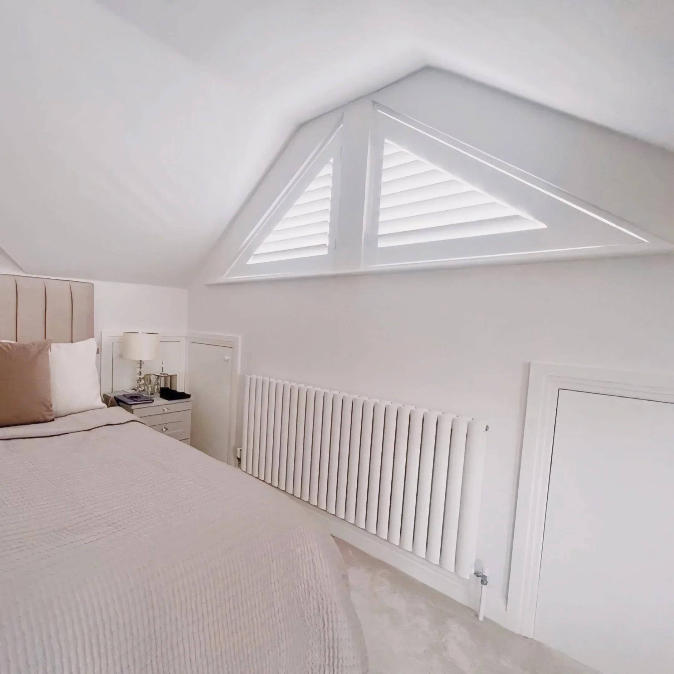 A white attic or loft room with a ceiling vent with white shutters, a bedside table with a lamp, books, and a beige cushion on a bed.