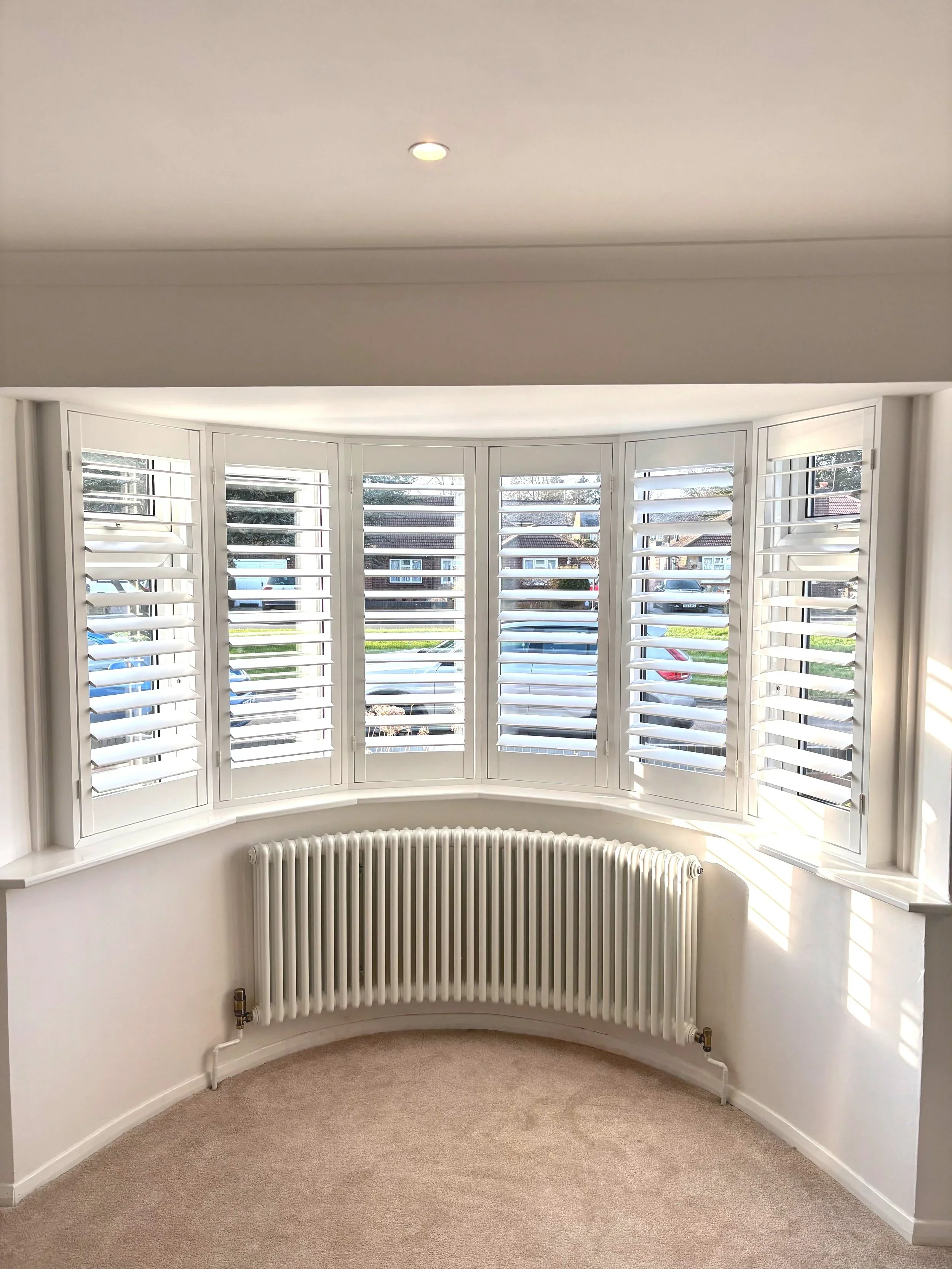 Room with large bay window featuring white plantation shutters, beige carpet, white walls, and a white radiator below the window.