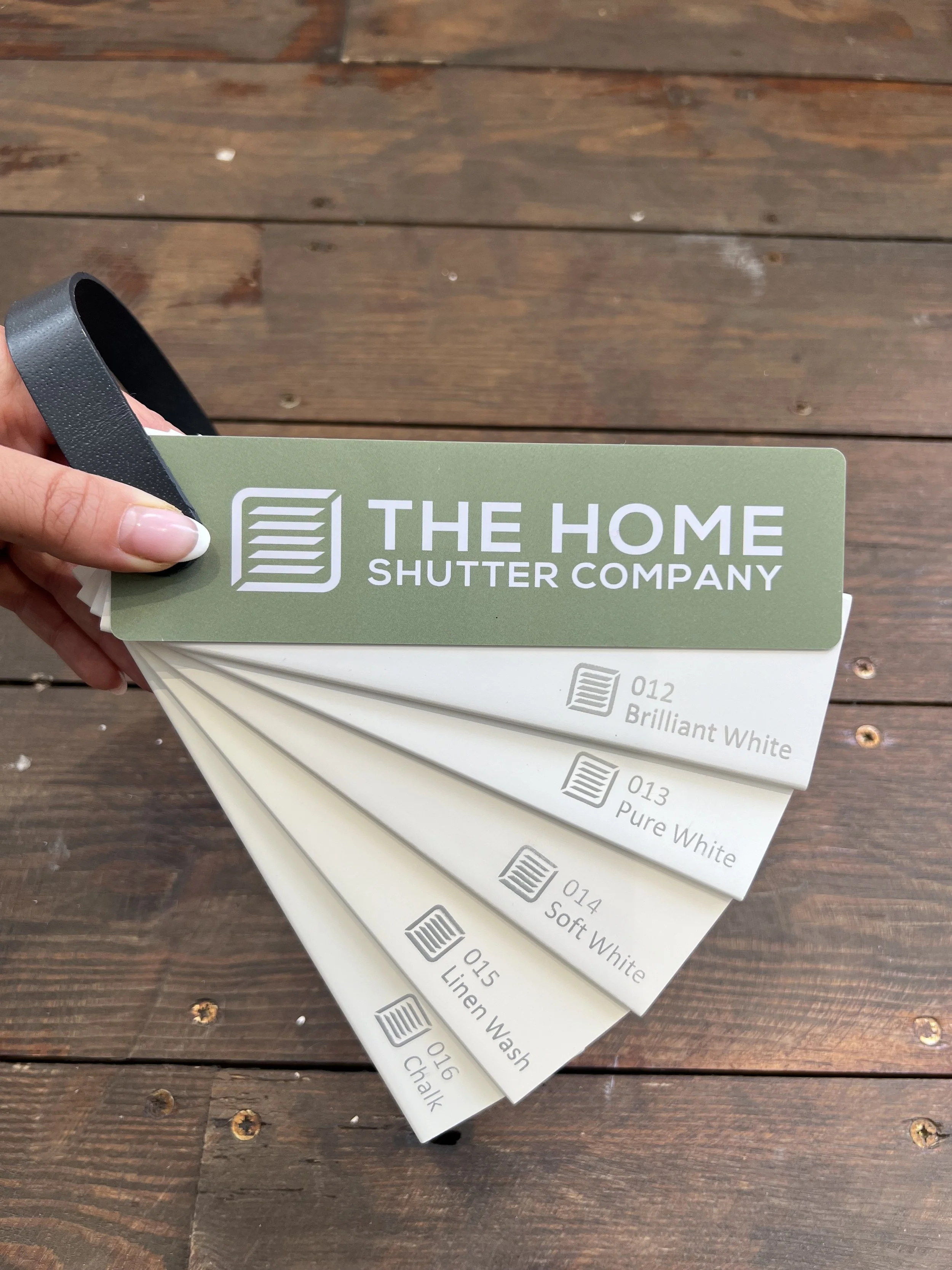 A person holding a set of paint color samples from The Home Shutter Company, fanned out over a wooden surface. The visible sample shows a color called 'Brilliant White' and others with different shades and names.