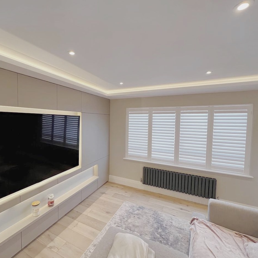 Modern living room with built-in entertainment center, large flat-screen TV, light-colored walls, hardwood flooring, a window with white blinds, and a gray radiator.