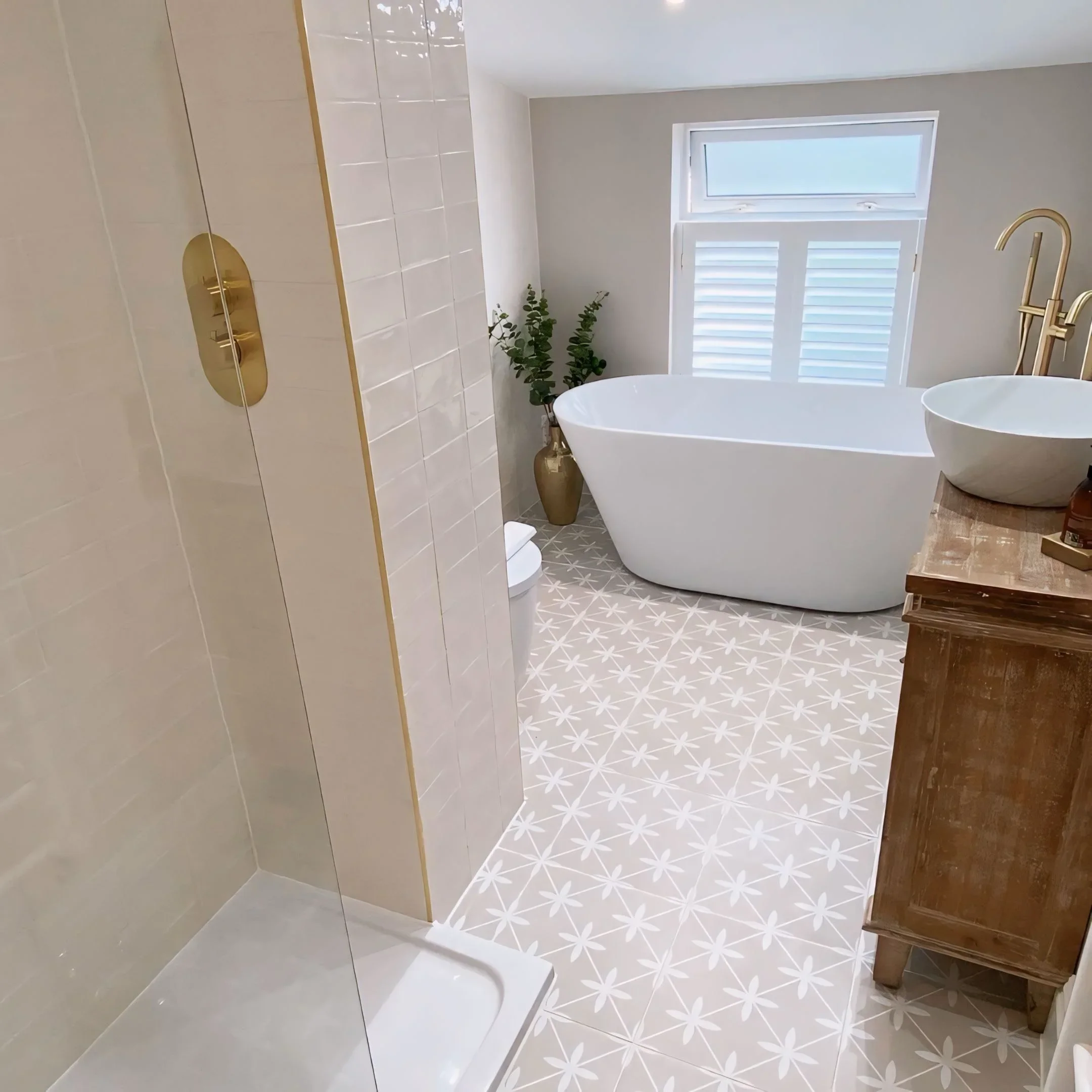 A modern bathroom featuring a white bathtub, a wooden side table, a golden faucet, a decorative plant, and a window with white blinds letting in natural light.