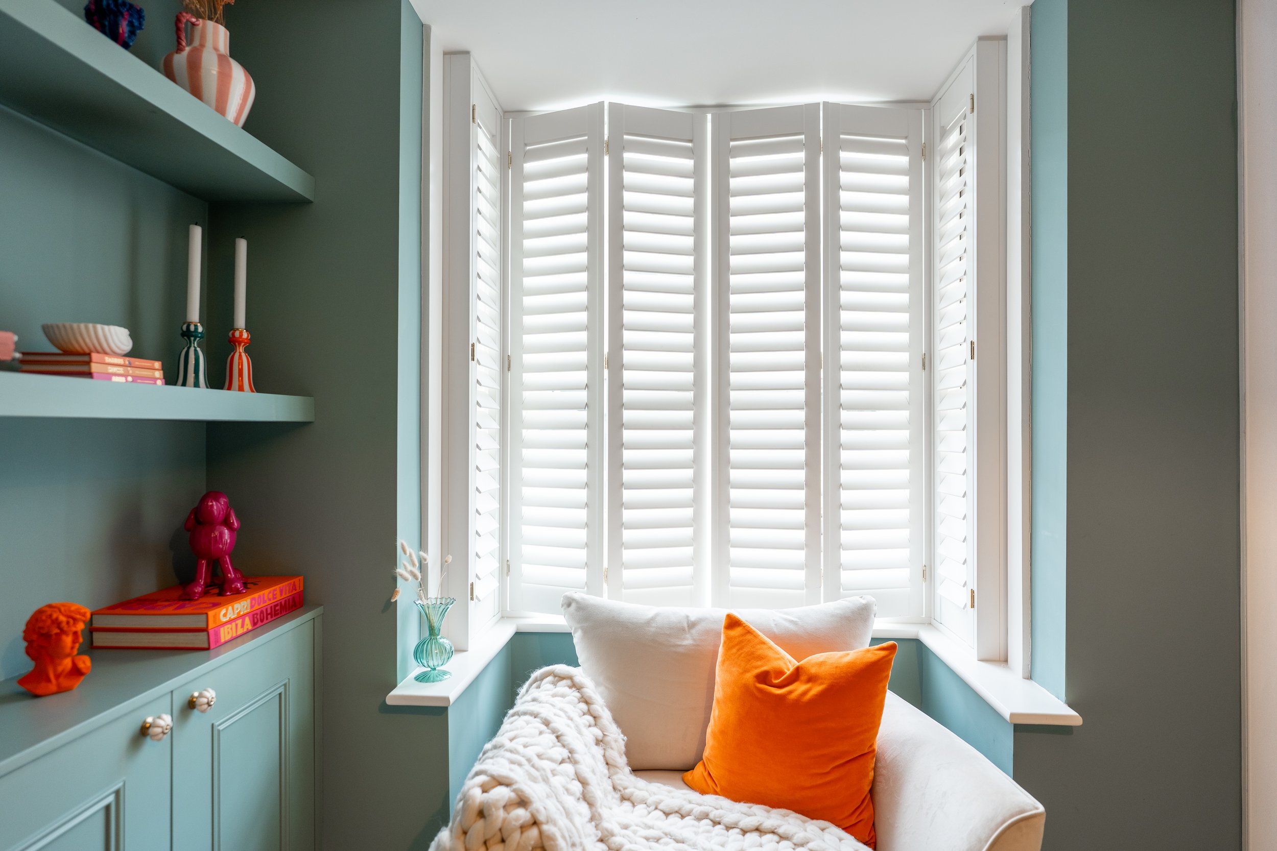 A cozy nook by a large bay window with white shutters, featuring a light turquoise built-in seating area with orange and white cushions, a small glass vase with white flowers, and a teal bookshelf with decorative items and books, including titles abo