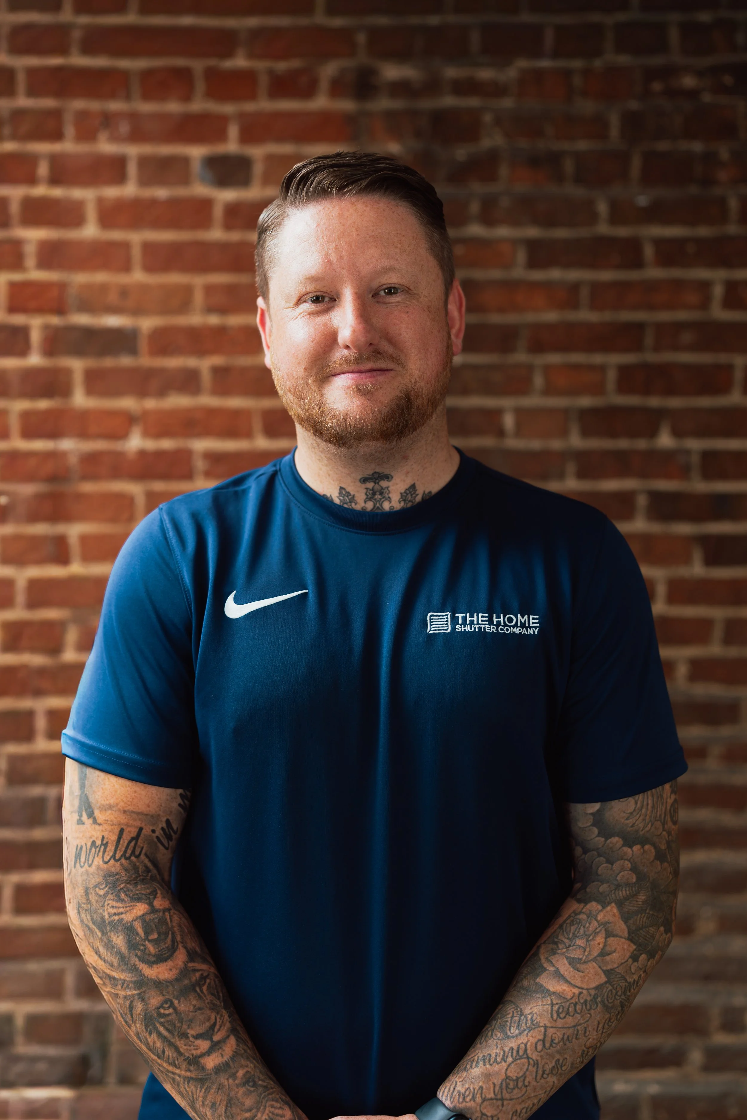 A man with short brown hair, a beard, and tattoos on his arms and neck, standing against a brick wall, wearing a blue Nike shirt with the logo and text 'The Home Shutter Company'.