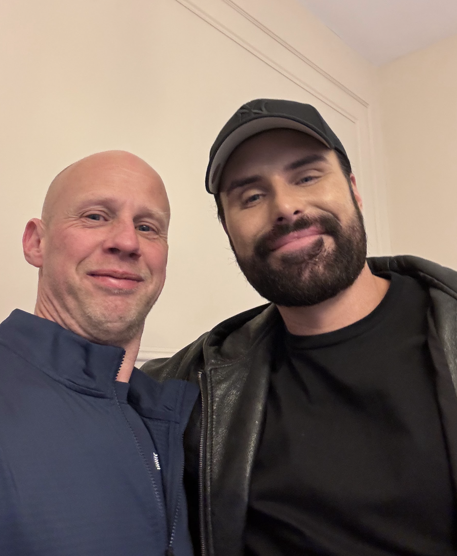 Two men taking a selfie. The man on the left is bald, wearing a navy blue jacket. The man on the right has a beard, wearing a black leather jacket and a baseball cap. They are indoors with a plain beige wall in the background.