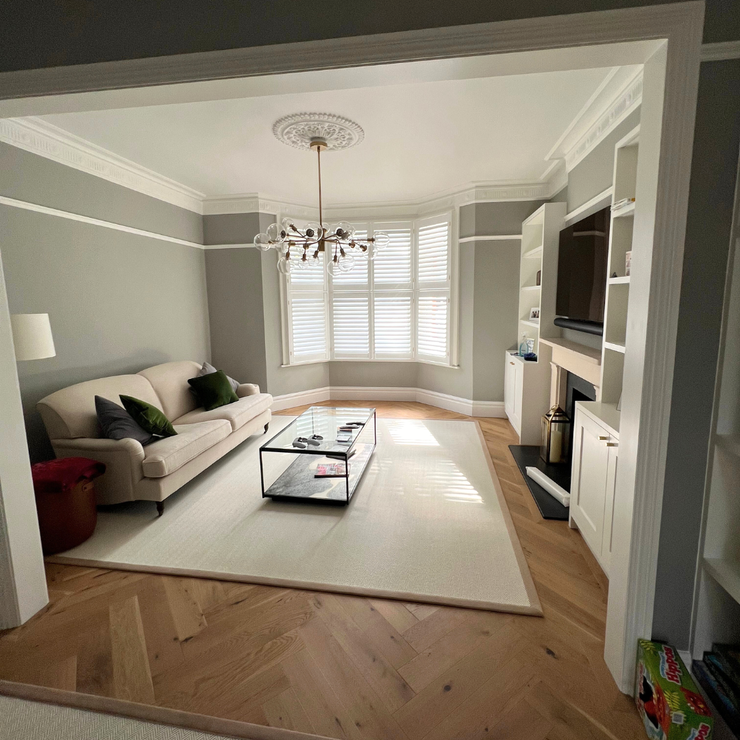 Living room with light green walls, white crown molding, a white sofa with dark and green pillows, a glass coffee table, a bay window with white shutters, a chandelier, and a fireplace with a TV above it. There is a white area rug and hardwood floors