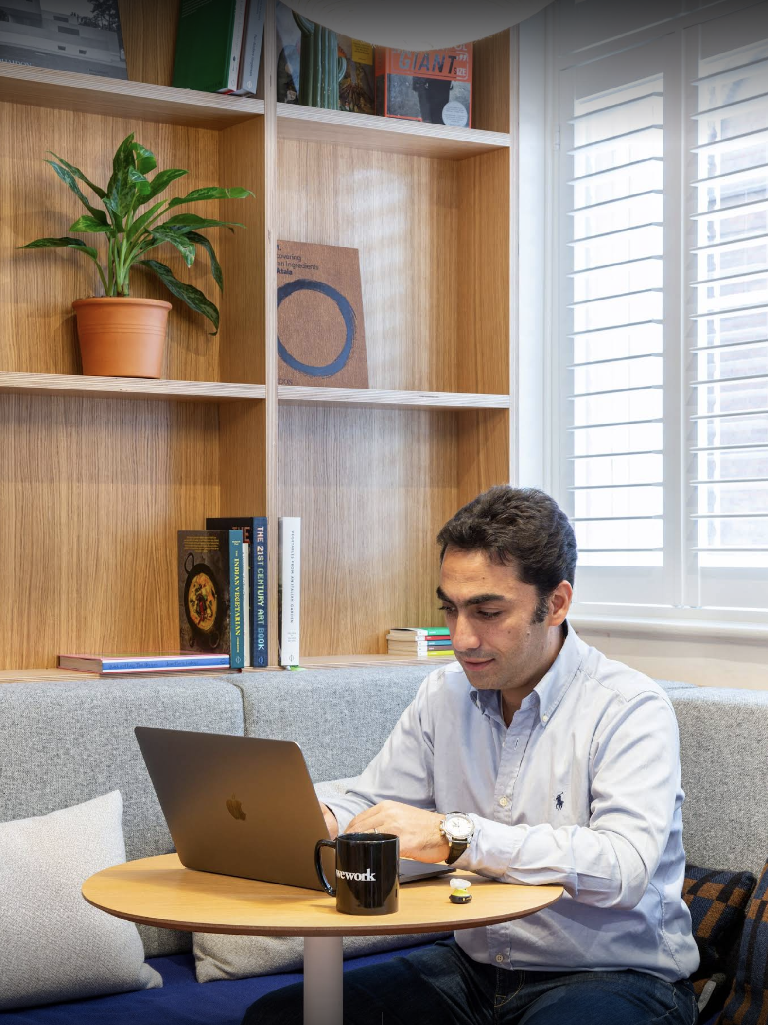 Man sitting at a round table working on a silver laptop in a cozy office setting with a bookshelf, window blinds, and a black mug on the table.