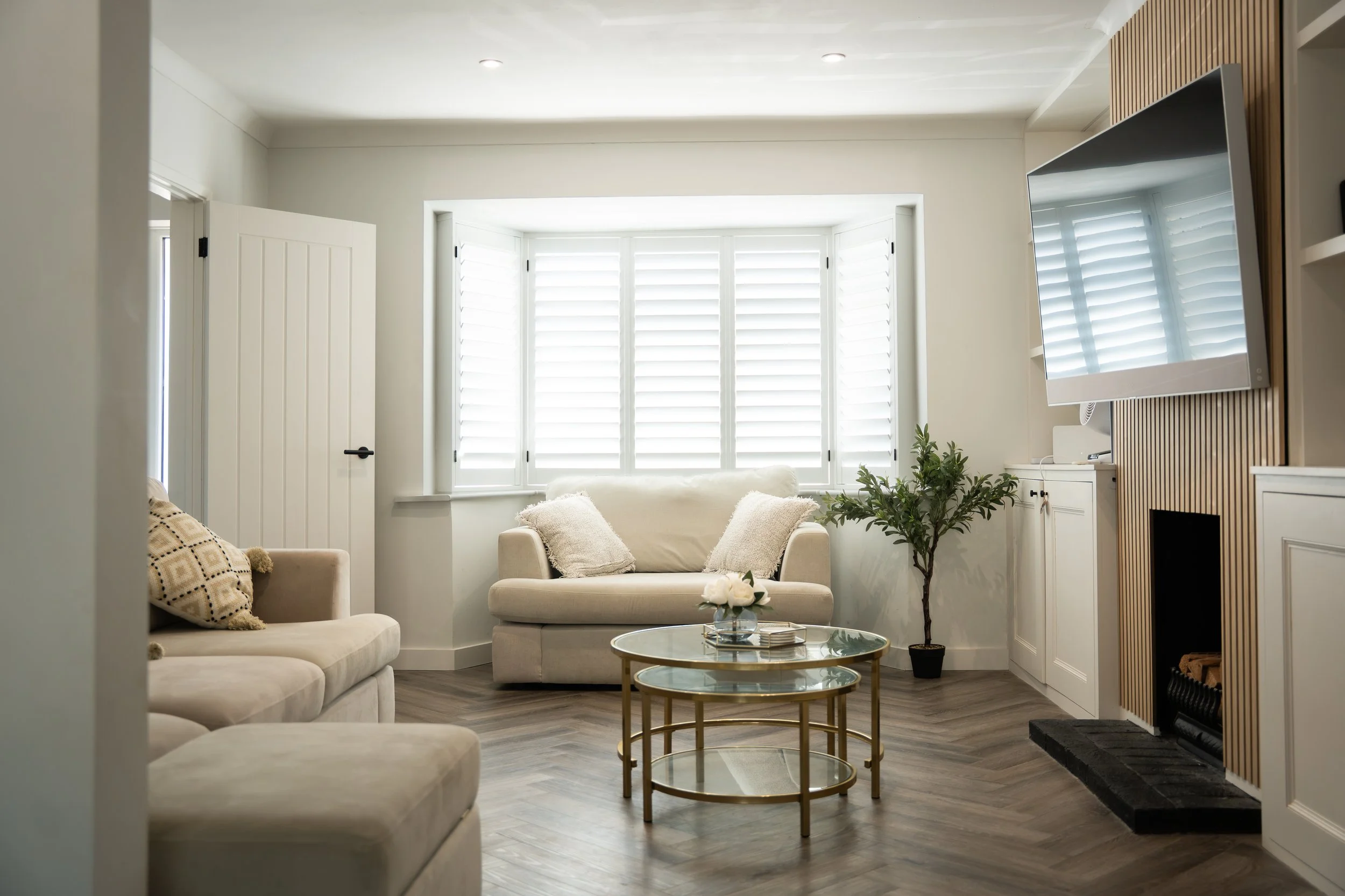 A cozy living room with beige sofas, a glass coffee table, a potted plant, a wall-mounted flat-screen TV, and large windows with white shutters.