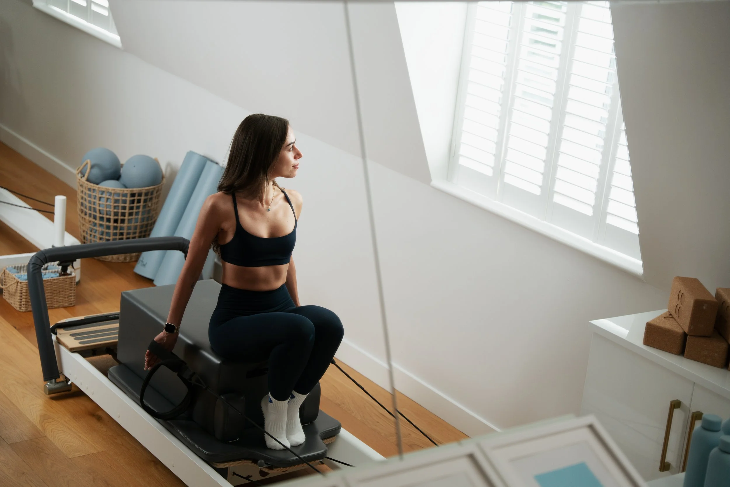 A woman sitting on a Pilates reformer machine in a bright room with large windows and white shutters, preparing for exercise.