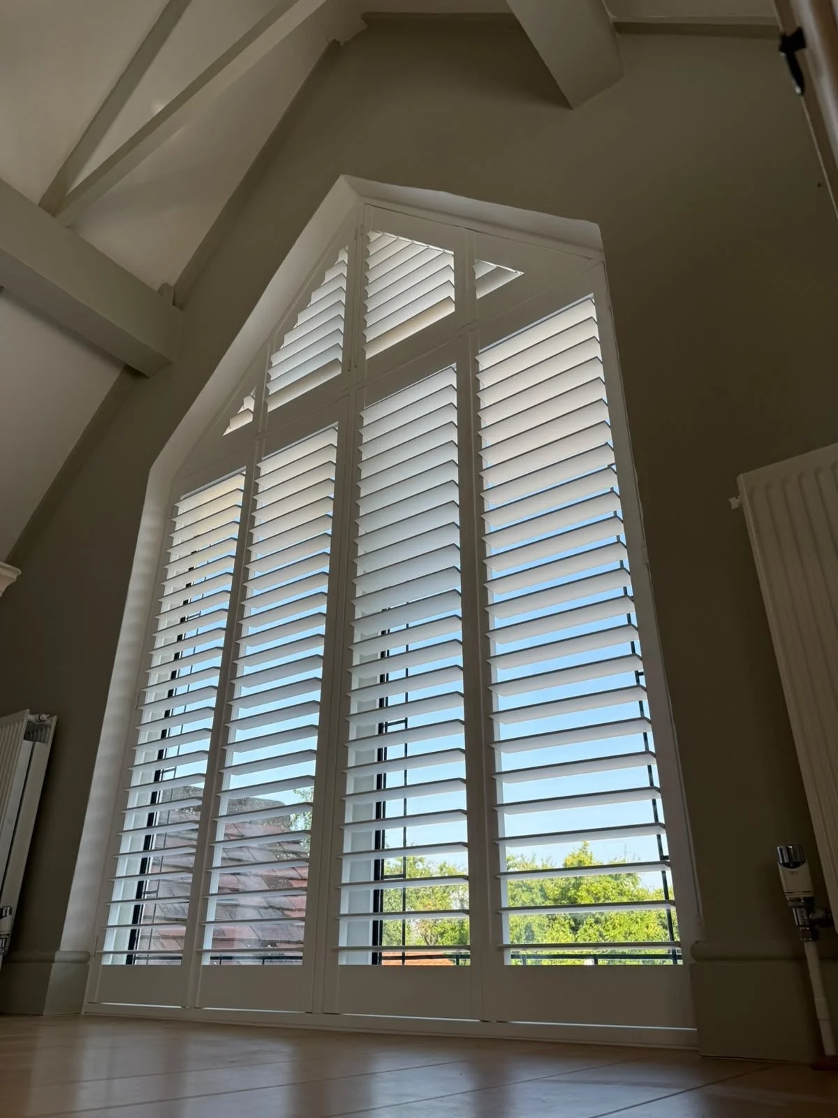A tall staircase window with white plantation shutters in a bright room, showing blue sky and trees outside.