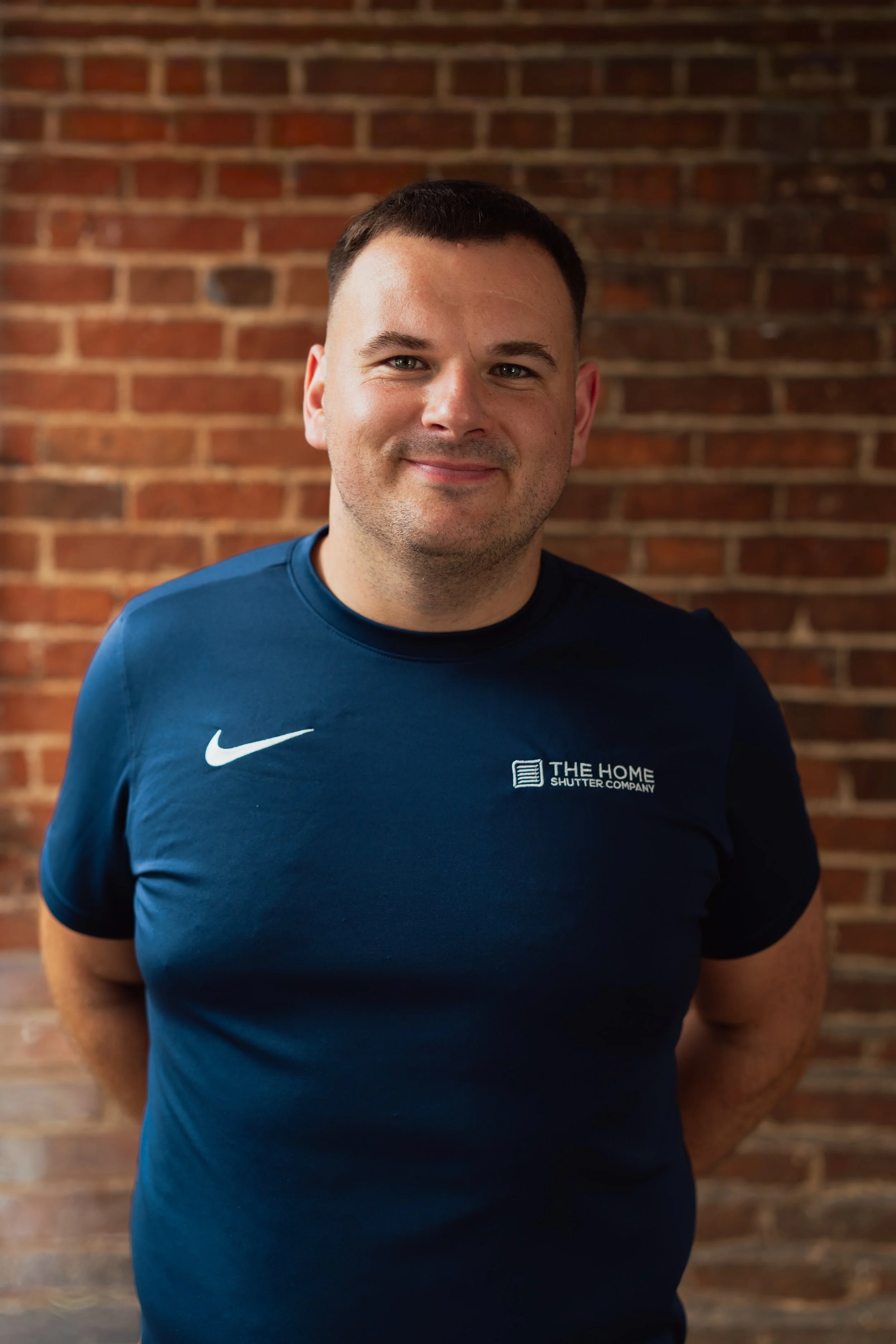 A smiling man in a blue Nike T-shirt standing in front of a brick wall.