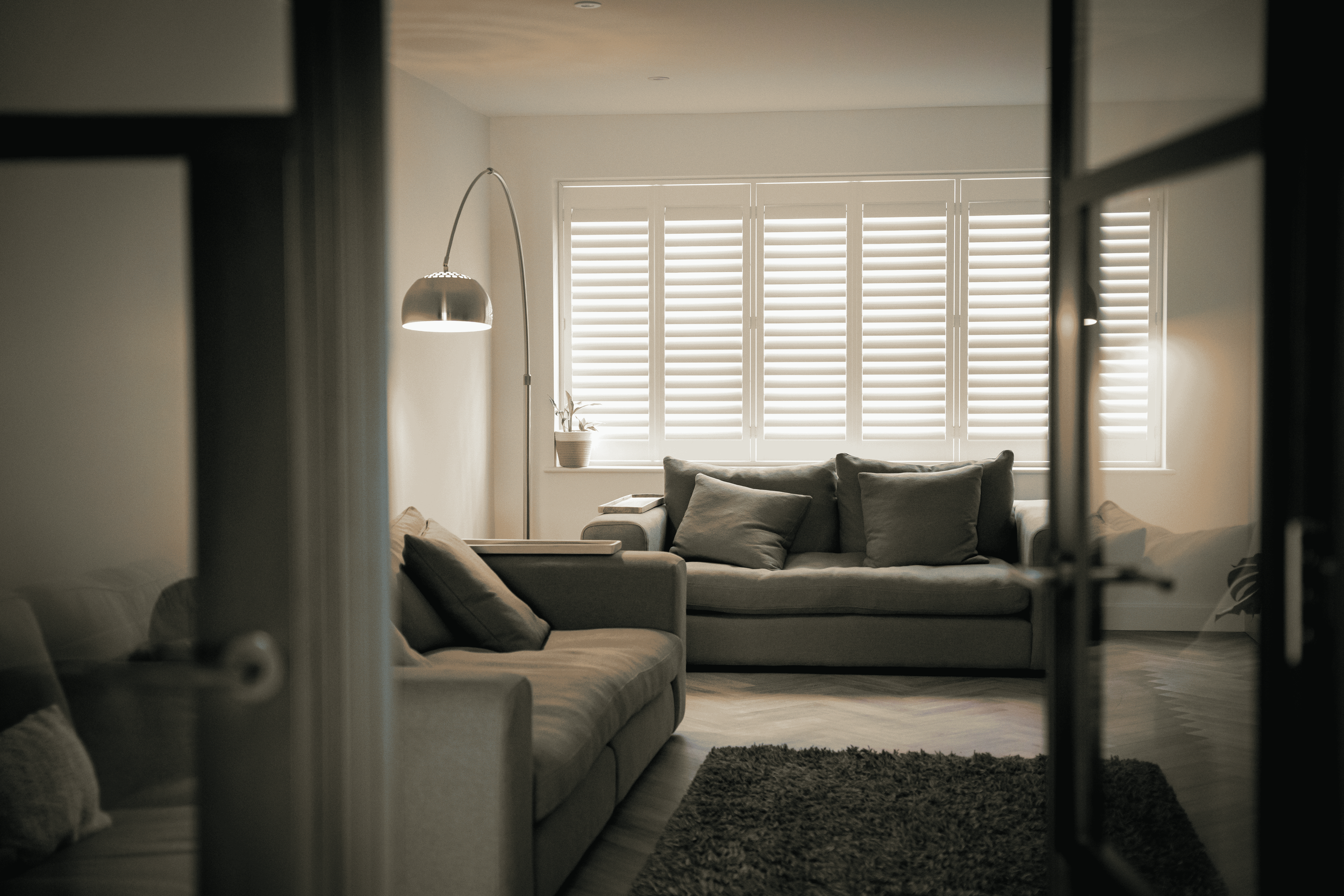 A cozy living room with a large window covered by white blinds, a gray sofa, a floor lamp, a potted plant, and a rug, viewed through a doorway.