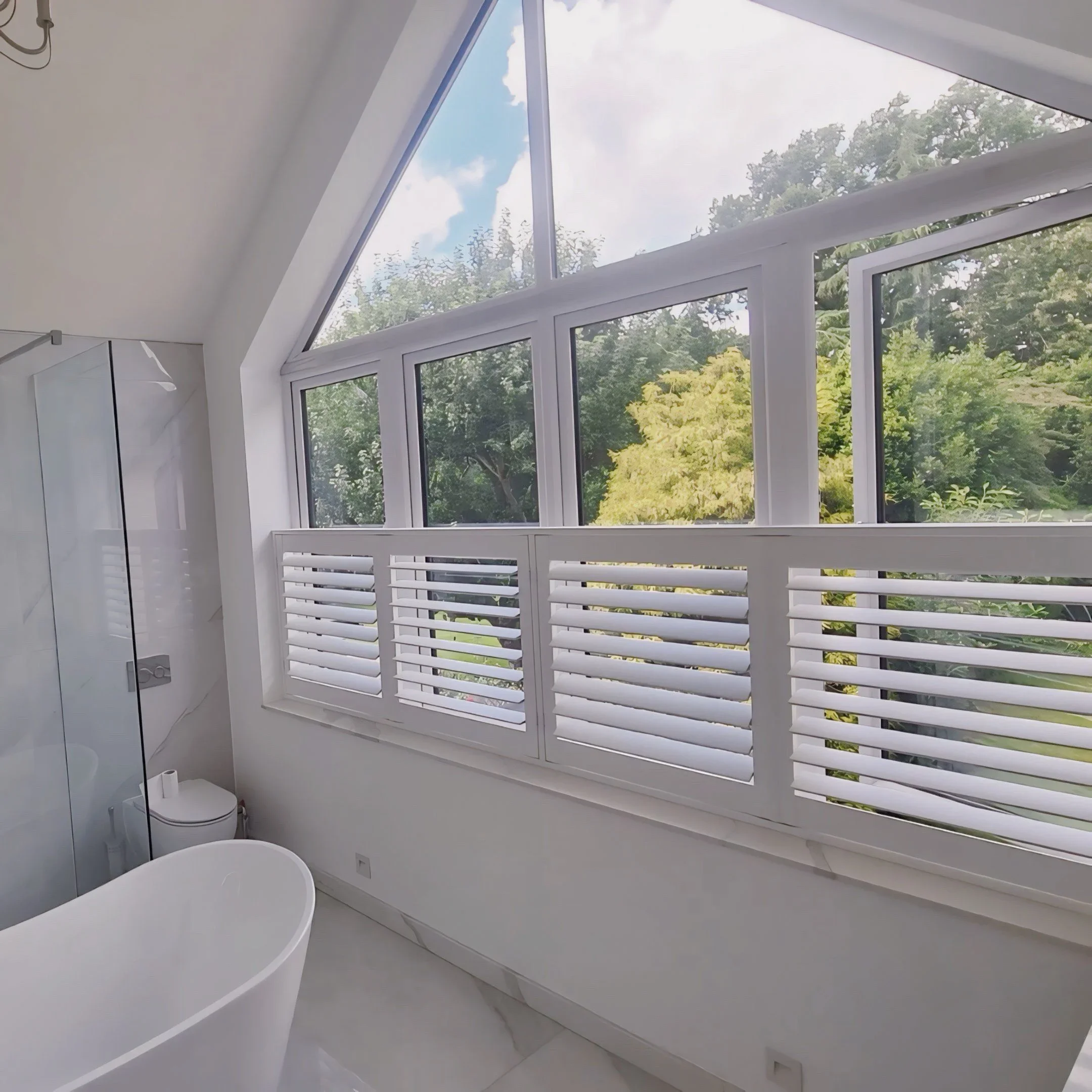 Interior view of a modern room with large white-framed windows overlooking green trees and a partly cloudy sky. The room has white walls, a white toilet, and white curtains.