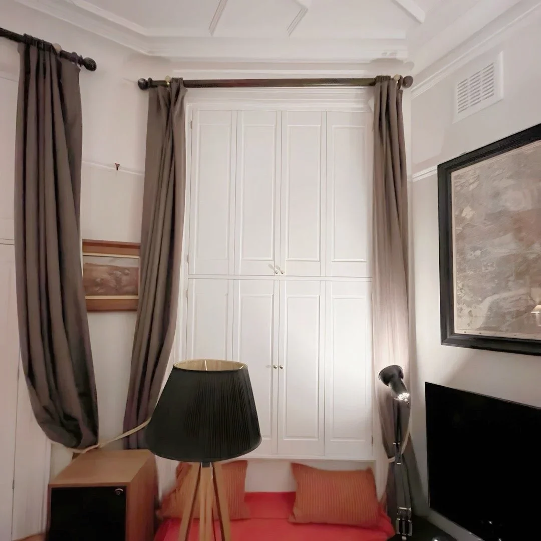 A cozy corner of a room with a bed or a daybed with orange pillows, a small wooden side table, a black lampshade on a tripod stand, dark curtains, a framed map on the wall, and white cabinets in the background.