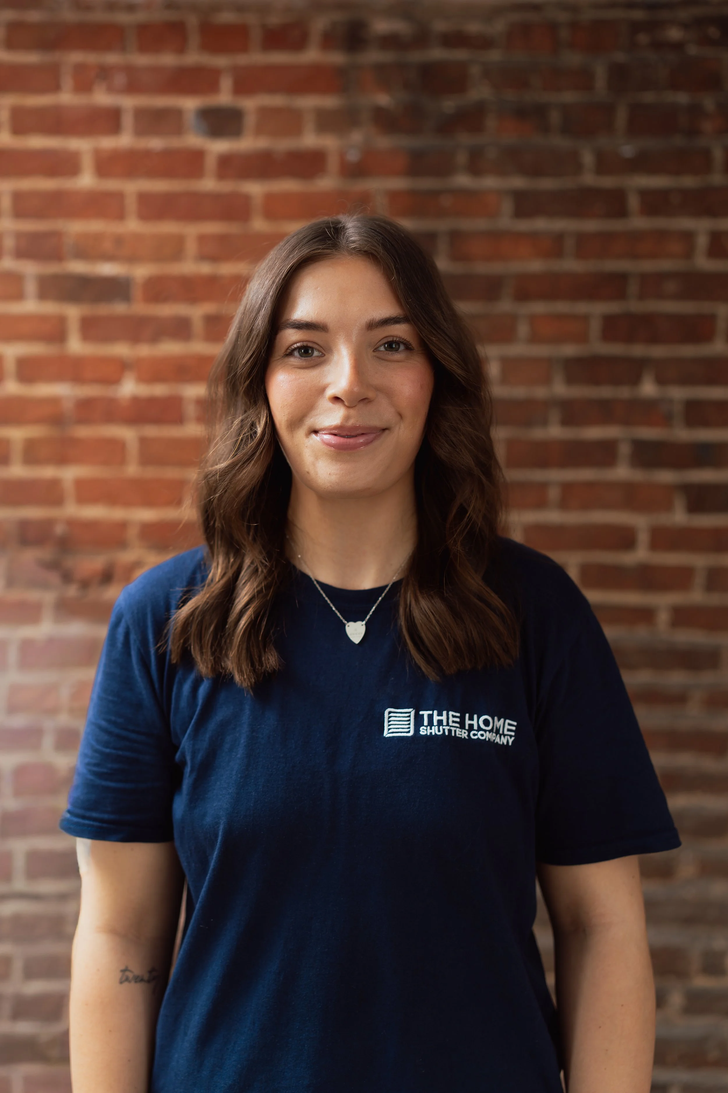 Portrait of a young woman with long brown wavy hair smiling, wearing a navy blue t-shirt with a company logo, standing in front of a brick wall.