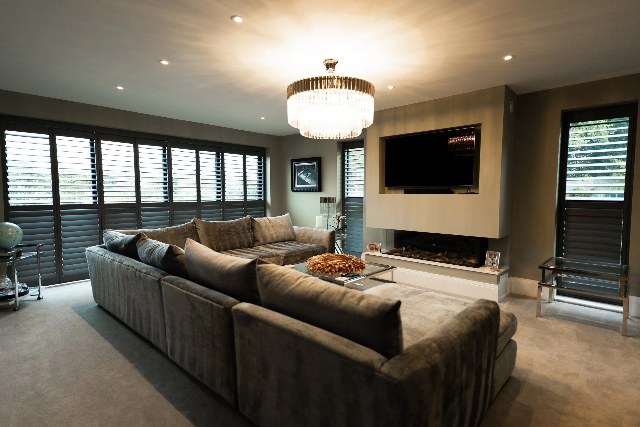 Modern living room with large sectional sofa, chandelier, flat-screen TV, and windows with plantation shutters