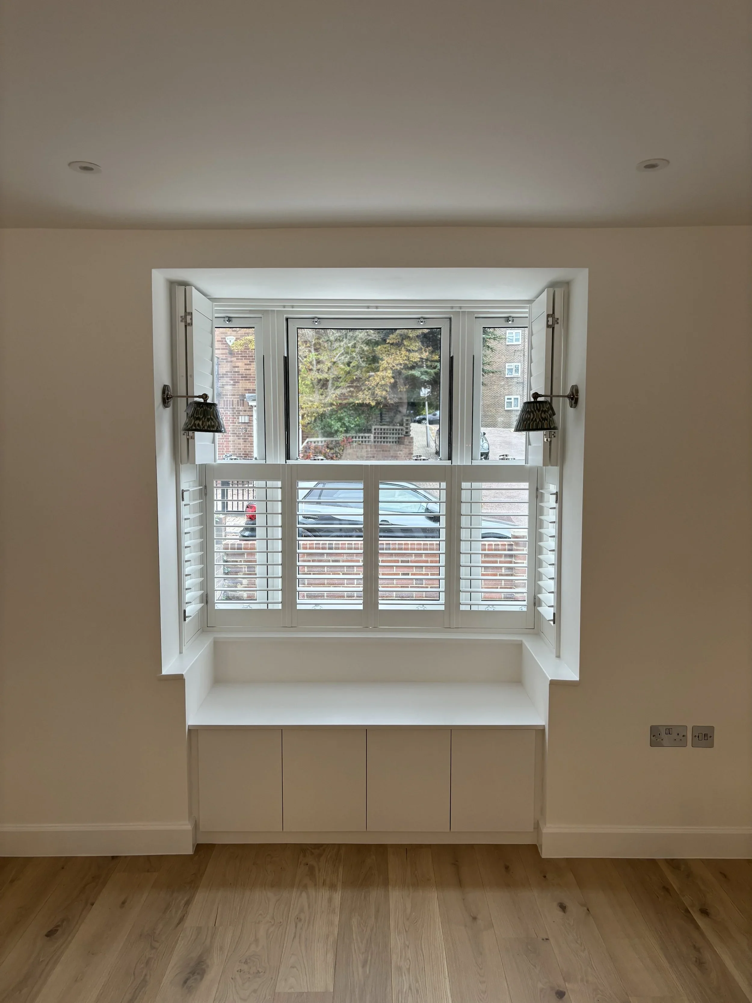 Interior view of a room with a large window featuring white plantation shutters, two wall-mounted lamps with patterned shades on either side, and a beige built-in cabinet below the window. Outside the window, trees and a brick building are visible.