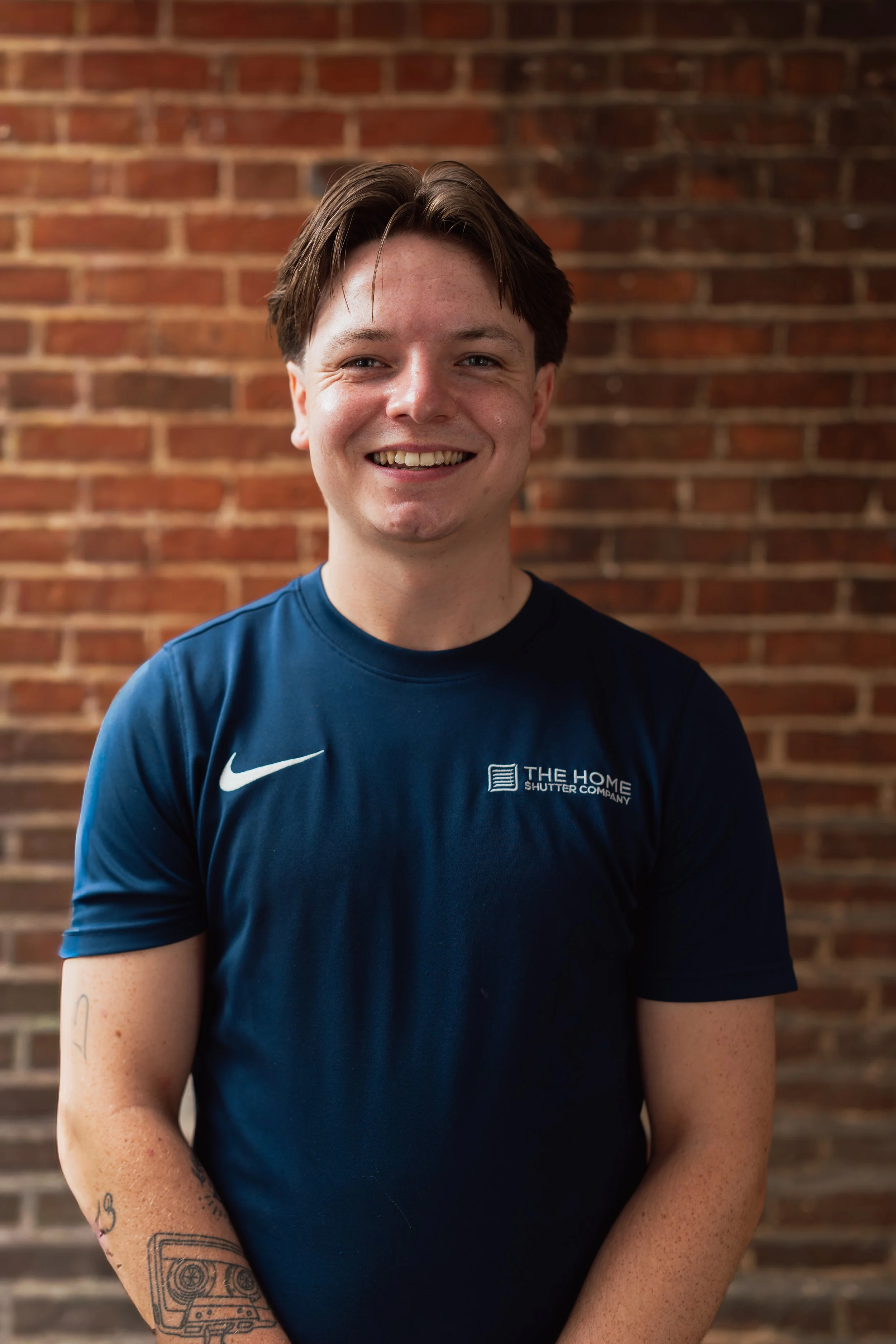 A young man with short brown hair, smiling, wearing a navy blue Nike T-shirt with a logo and text on it, standing against a brick wall background.