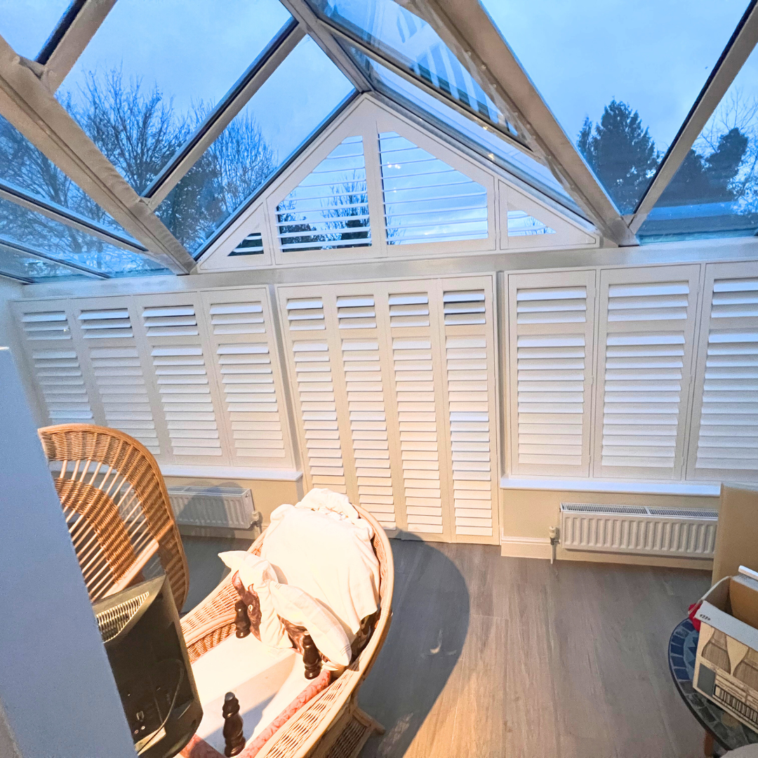 An enclosed porch or sunroom with large glass windows and a triangular glass ceiling. There is a wicker daybed with a blanket and a pillow. A box is on a small round table, and a radiator is mounted on the wall beneath the windows. Outside trees are 