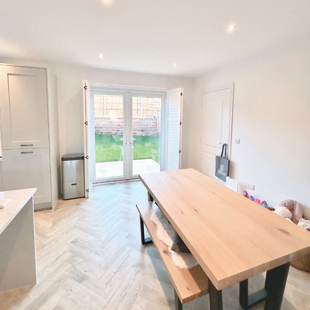 Bright dining room with a large wooden table, a bench, and chairs, open to a backyard with a patio, grass, and a wooden fence, with white walls and ceiling, and a sliding glass door with shutters.