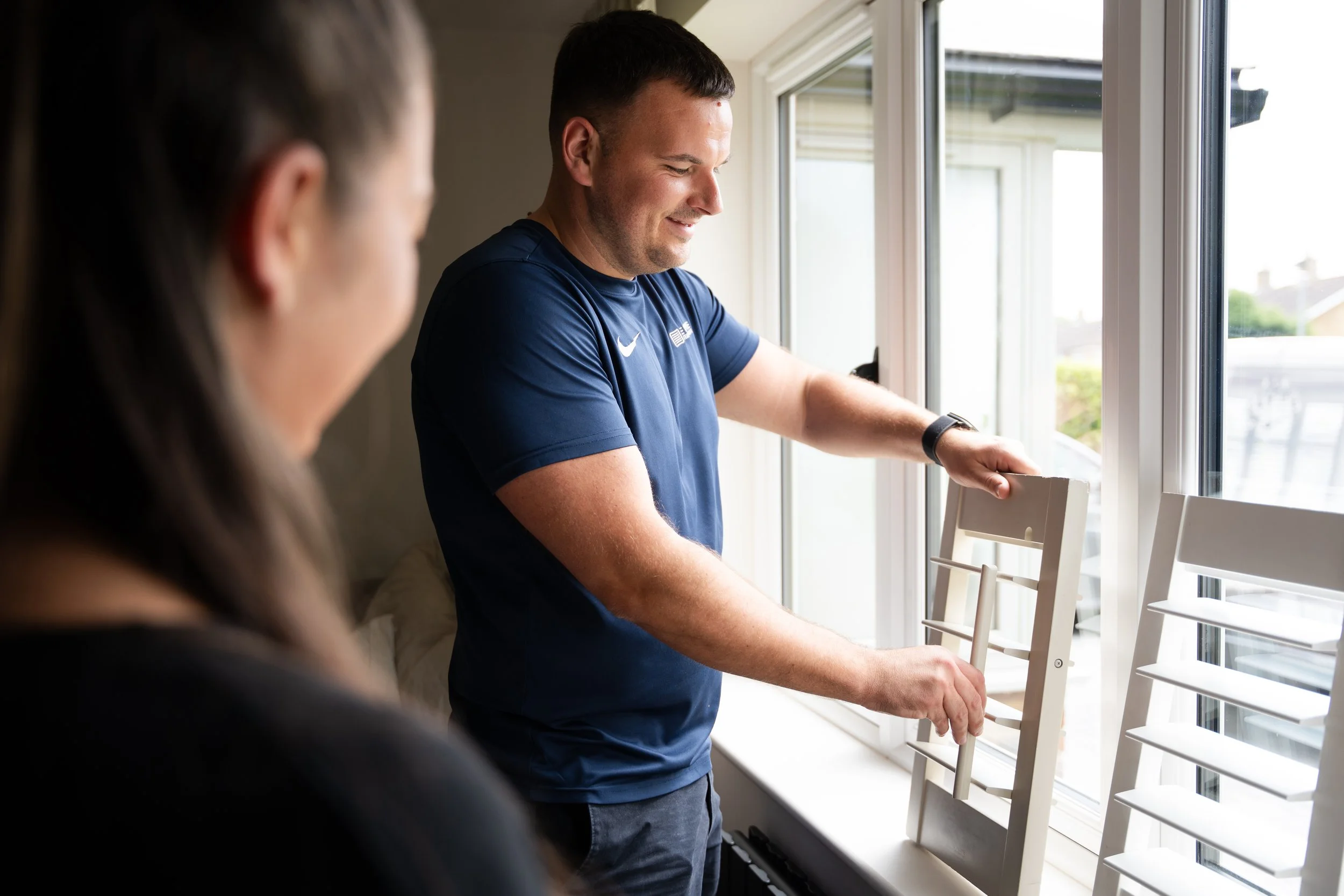 A man wearing a navy blue shirt and a woman are standing near a window with white blinds, with the man holding a window shutter slat in his hands, possibly adjusting or installing it.