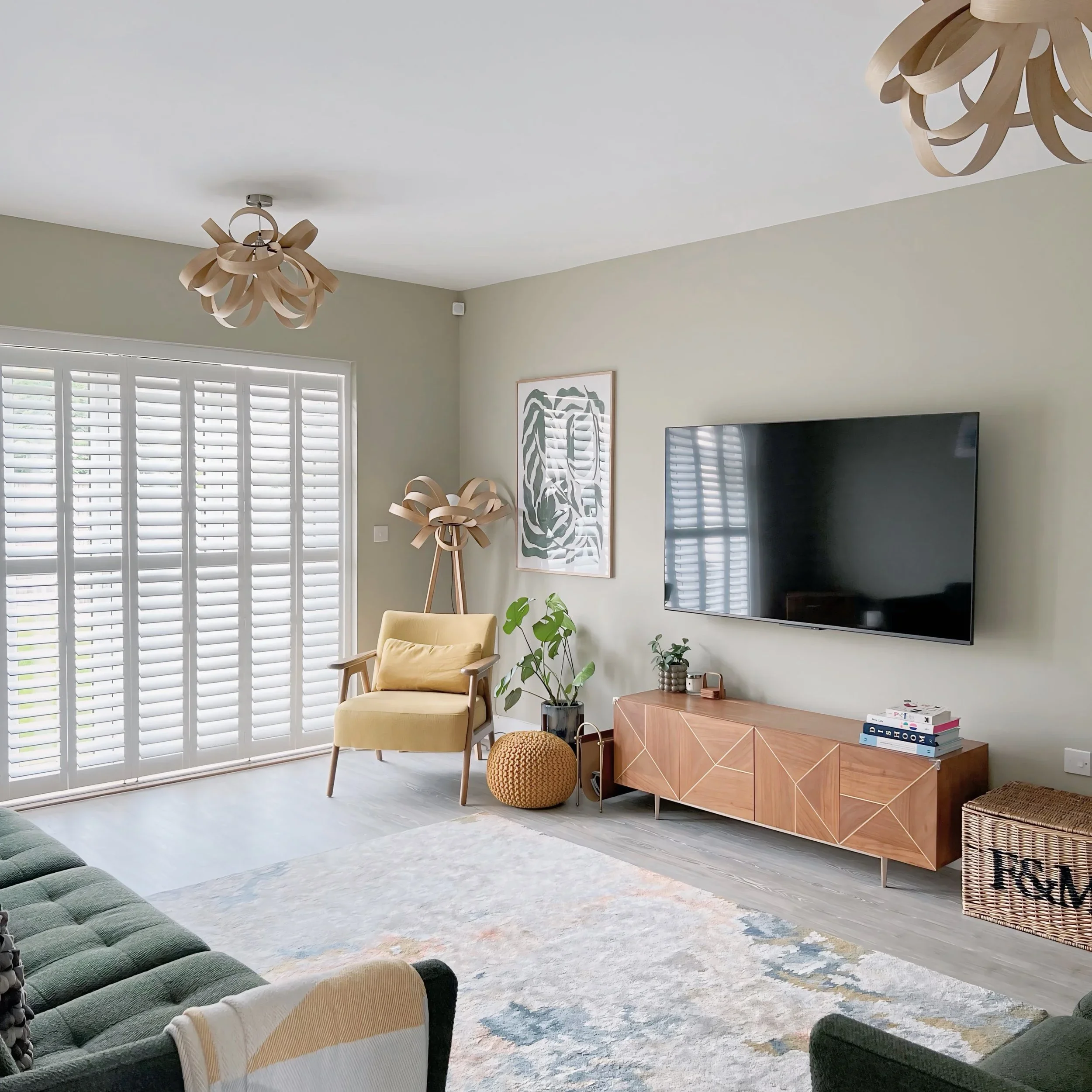 A living room with a green sofa, a yellow armchair, a wooden sideboard, a large flat-screen TV, multiple plants, artwork on the wall, window shutters, and modern light fixtures.