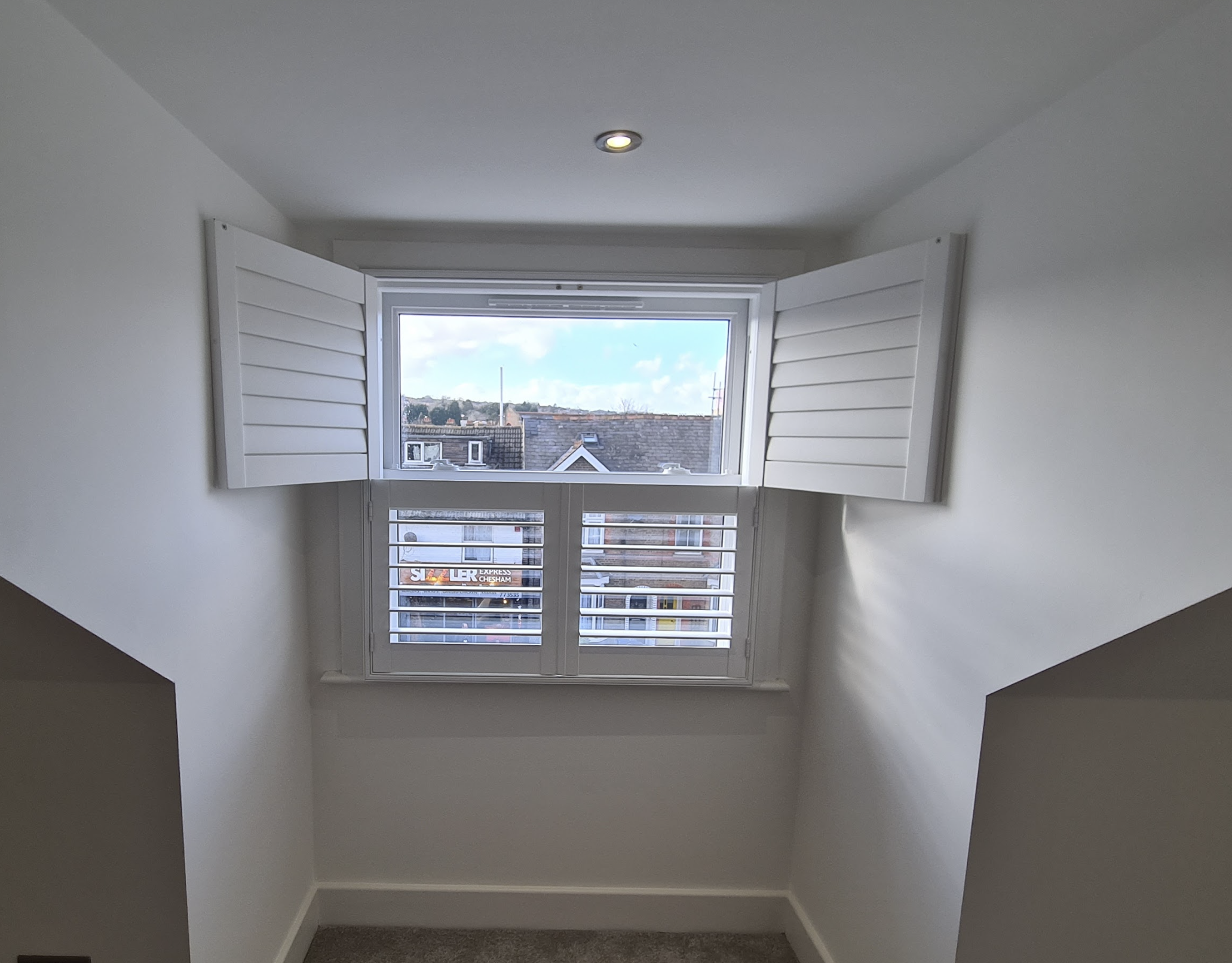 An attic room with an open window showing a view of rooftops and a cloudy sky, with white walls and gray carpeted floor.